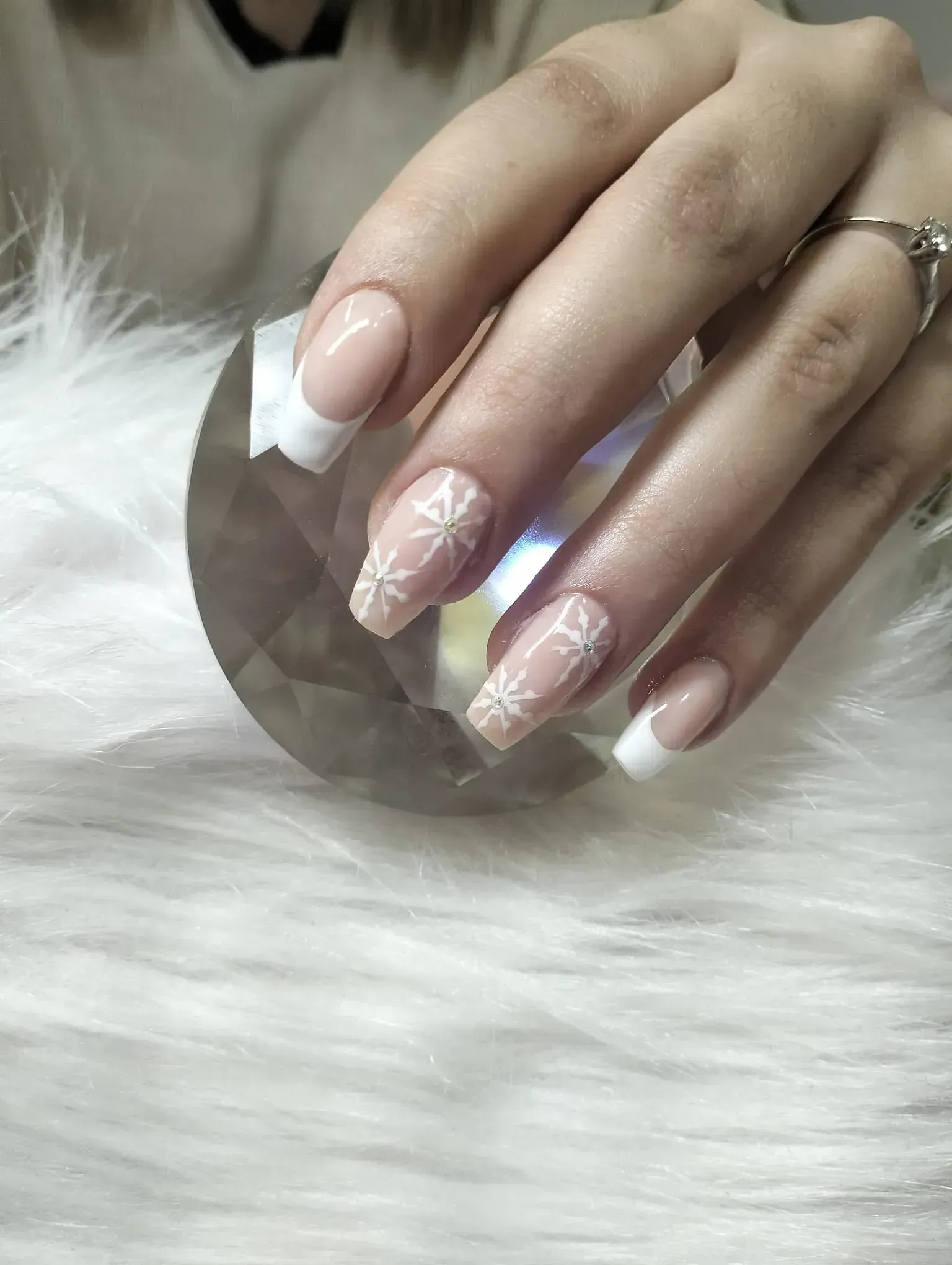 Mano con punta francesa y uñas con diseño de copo de nieve sobre un cristal, apoyada sobre un material blanco esponjoso.