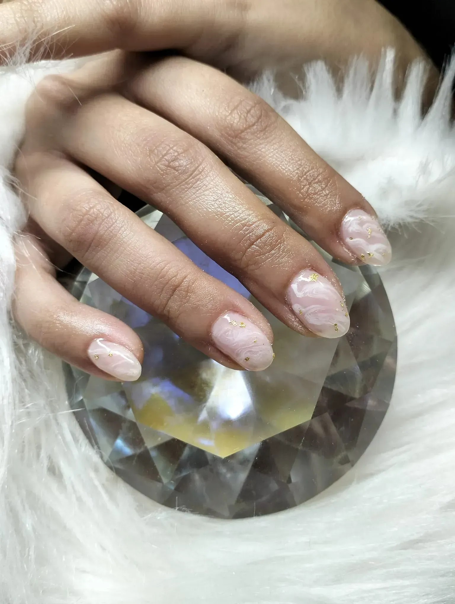 Mano con uñas de forma ovalada, efecto mármol, apoyada sobre un cristal transparente, con pelaje blanco de fondo.