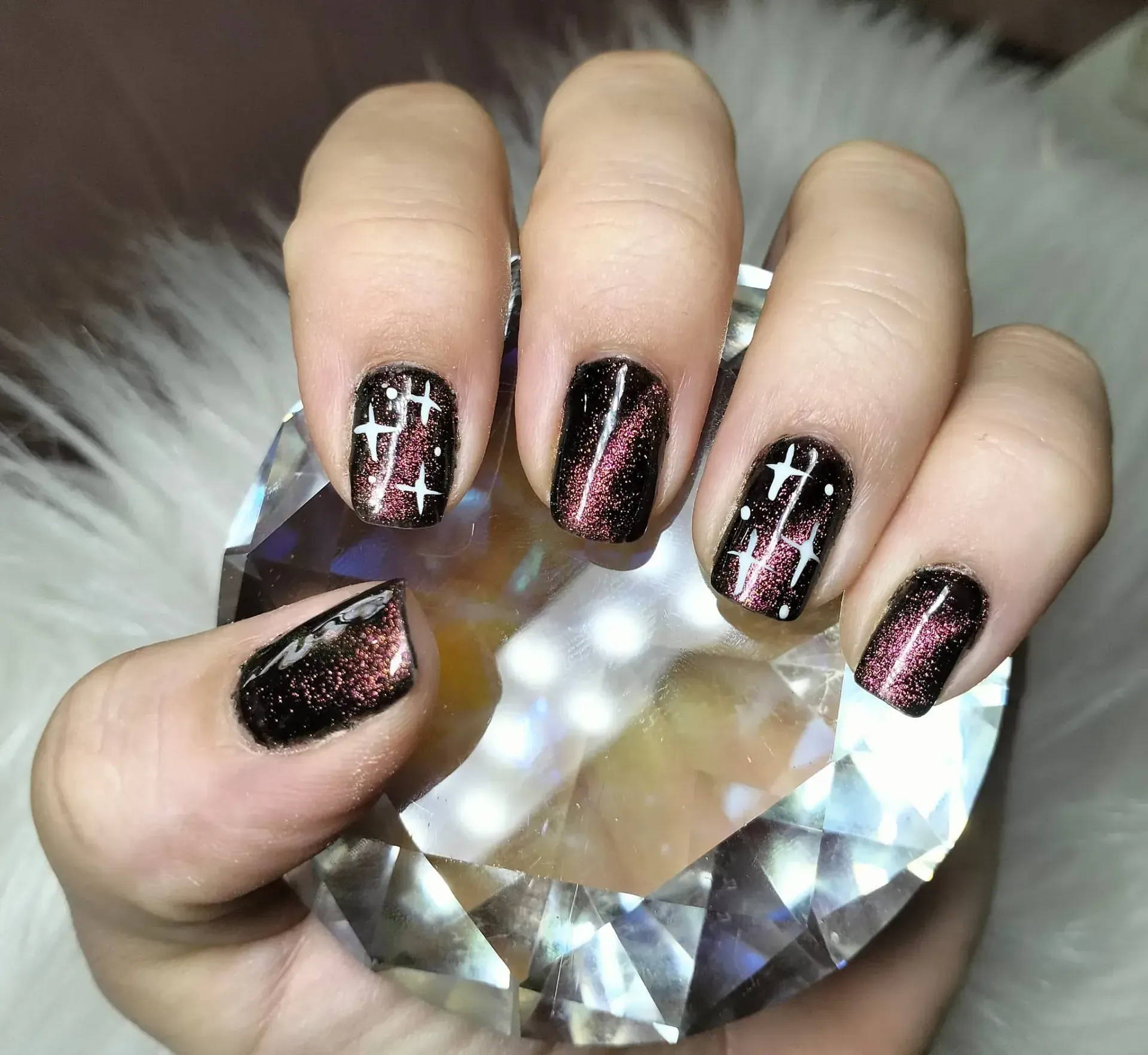Mano con uñas negras, brillo rojo y diseño de cruz blanca, sosteniendo un cristal.
