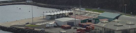 Vista aérea de uma instalação portuária com vários edifícios, píeres e barcos, com uma floresta ao fundo.