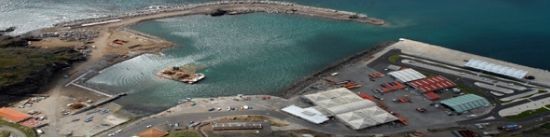 Vista aérea de um porto com prédios, um píer e água ao redor. Os prédios têm telhados brancos, vermelhos e verdes.