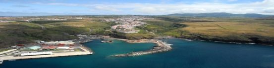Vista aérea de uma cidade costeira aninhada entre colinas verdes e uma baía azul-turquesa. Um porto se estende até a água.