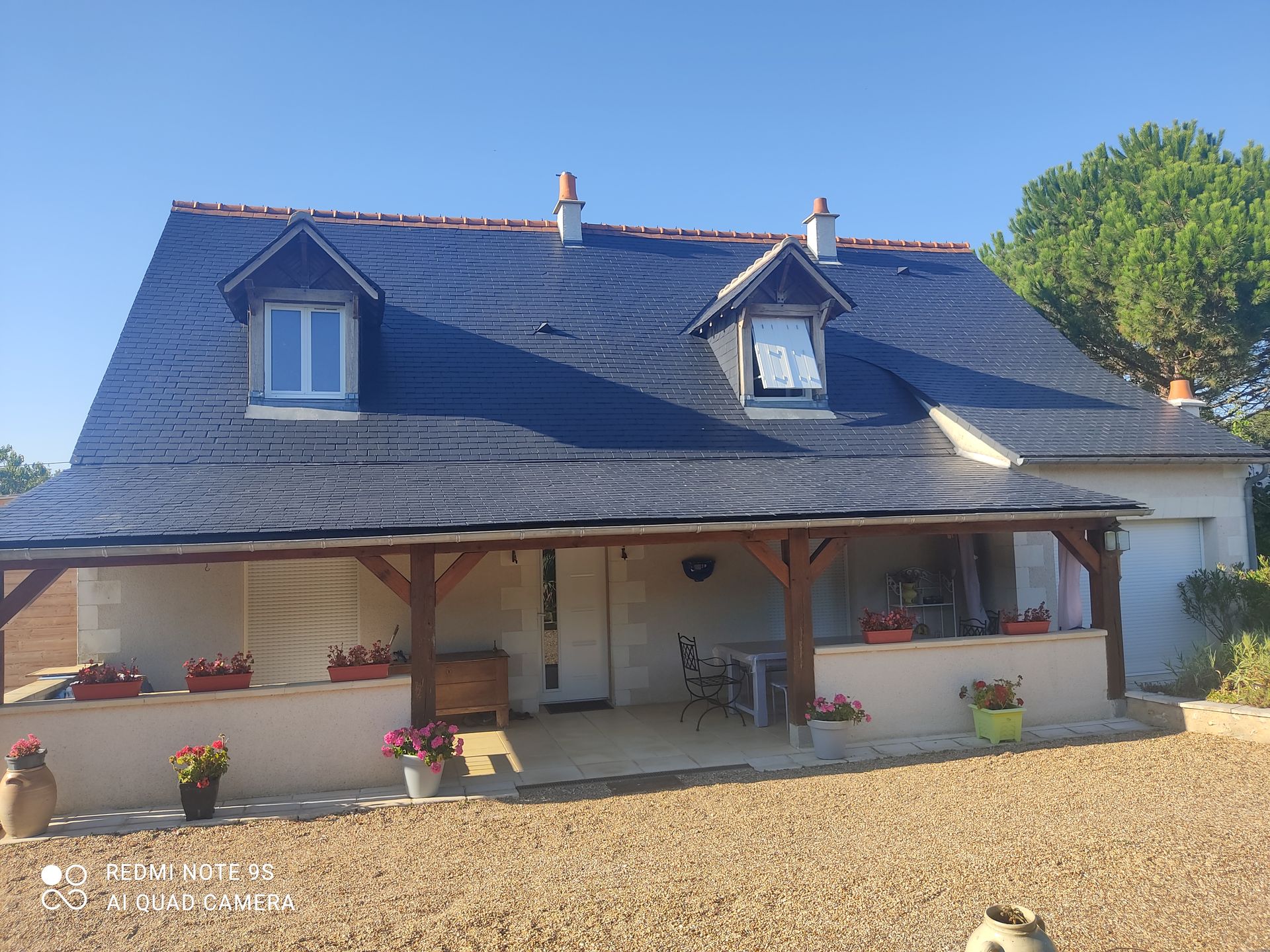 Maison au toit sombre avec deux lucarnes, porche, cour gravillonnée et ciel bleu azur.
