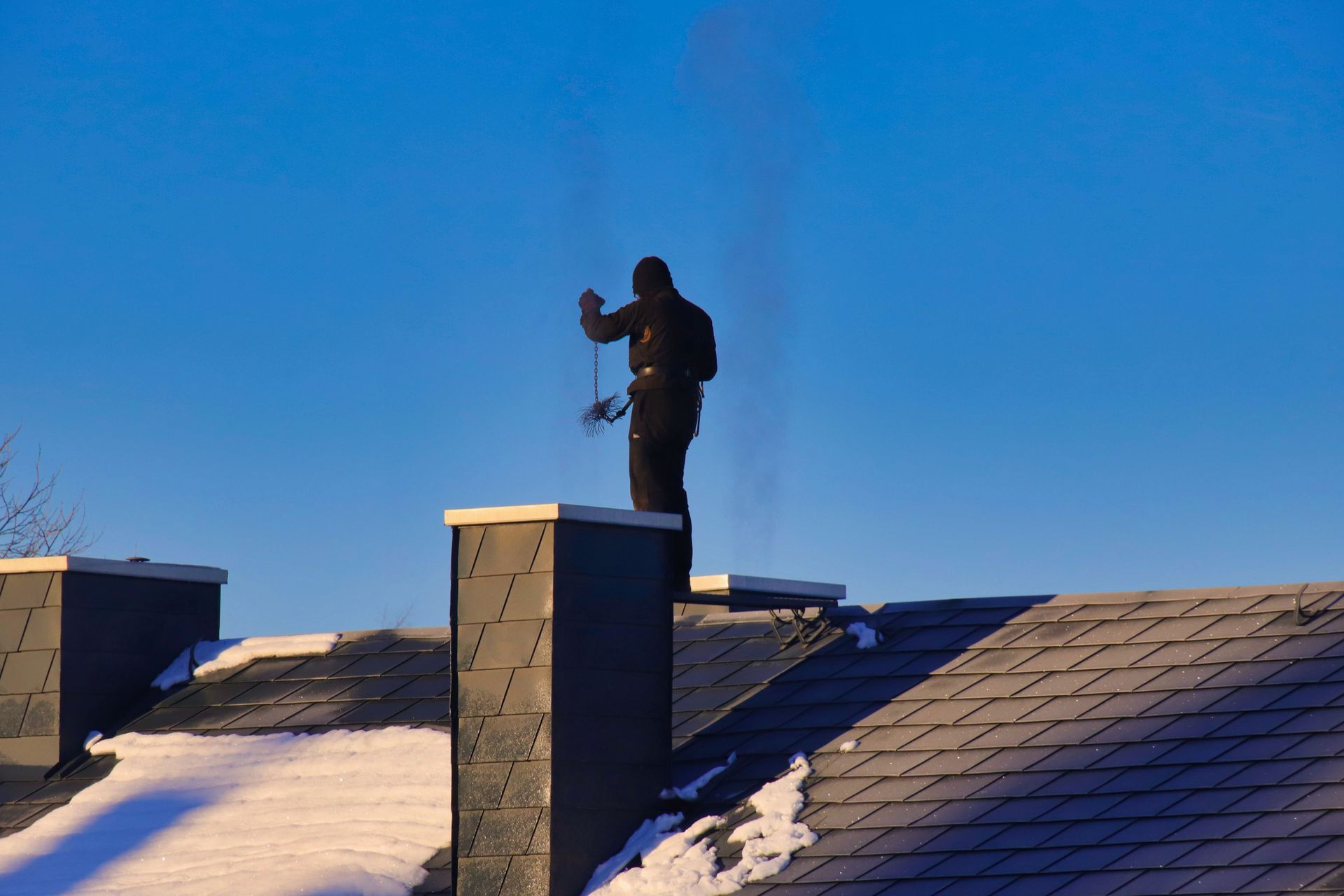Un ramoneur nettoie une cheminée sur un toit, devant un ciel bleu parsemé de neige.