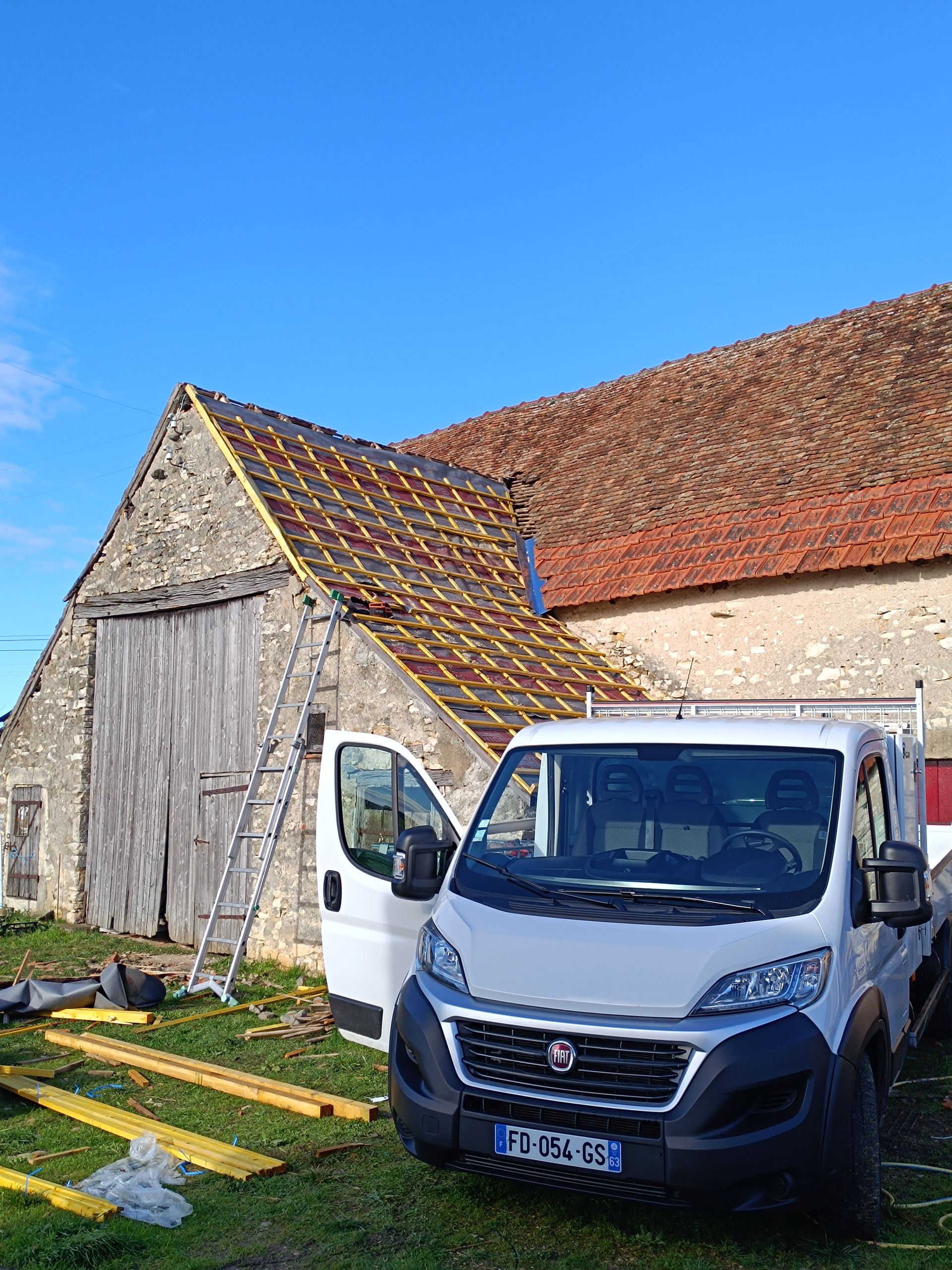 Une camionnette blanche est garée près d'une grange au toit de tuiles. Une échelle est appuyée contre la grange. Ciel bleu.