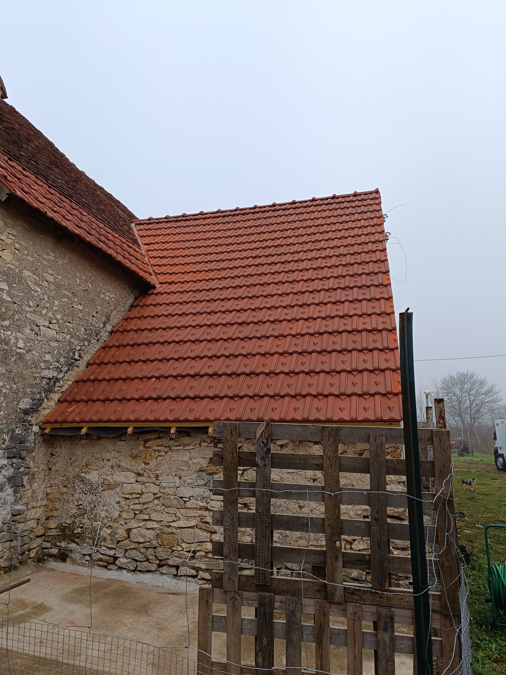 Toit de tuiles rouges sur un vieux bâtiment en pierre avec une clôture en bois devant, journée nuageuse.