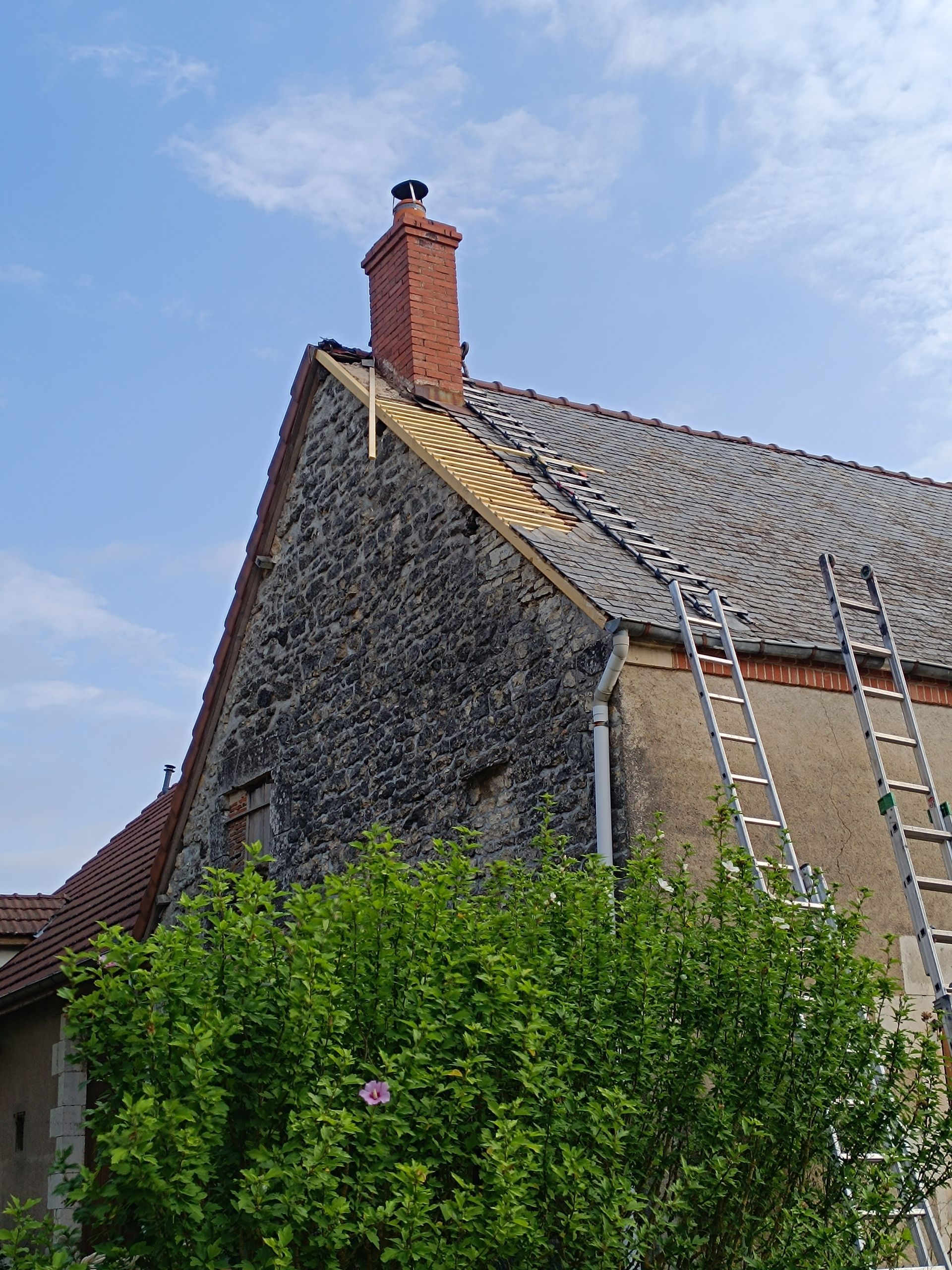 Une cheminée en briques surplombe un toit dont il manque des tuiles. Une échelle est appuyée contre le mur de la maison.