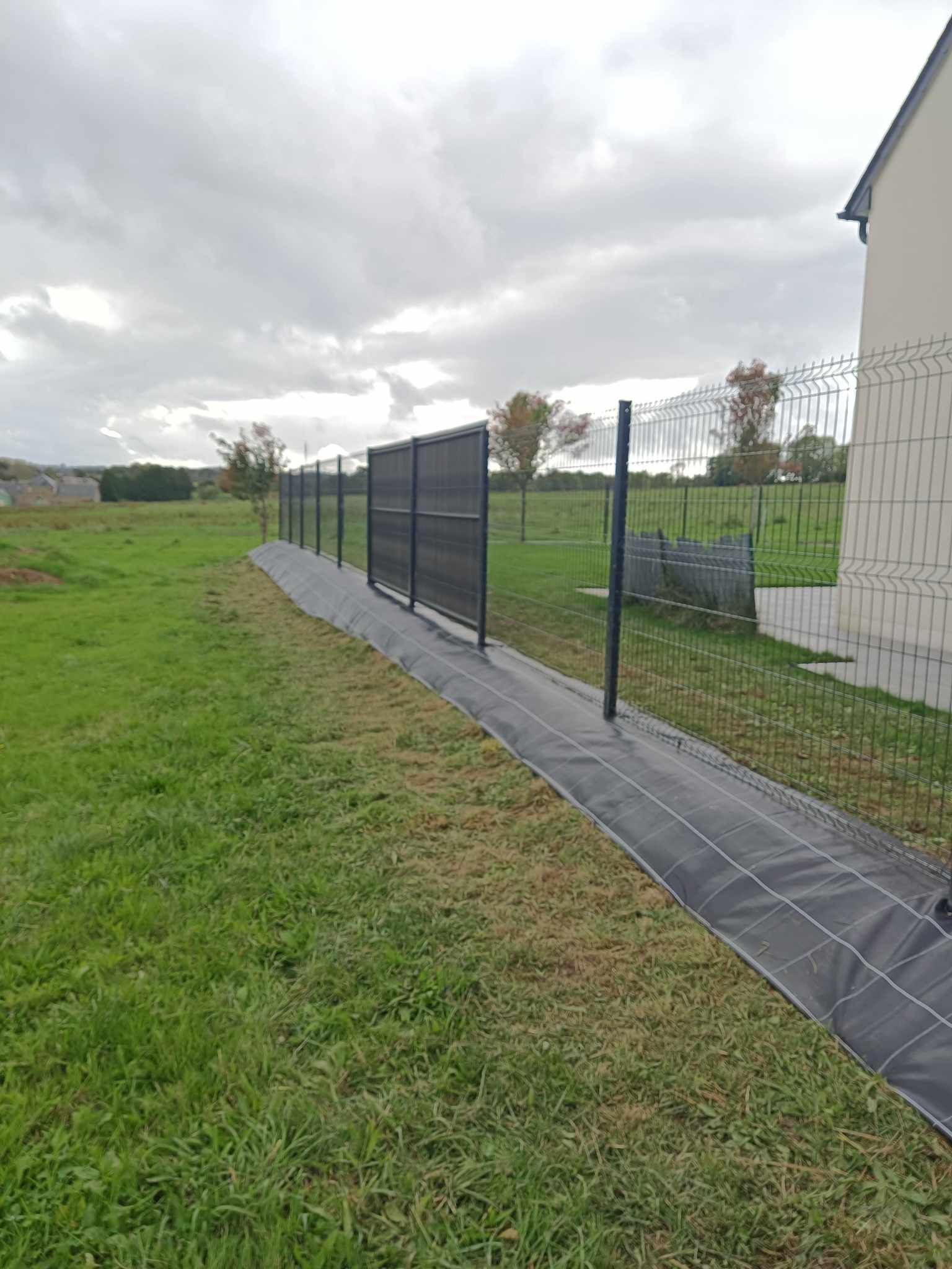 Clôture en grillage noir le long d’un chemin pavé, à côté d’un champ enherbé et d’un bâtiment blanc, sous un ciel nuageux