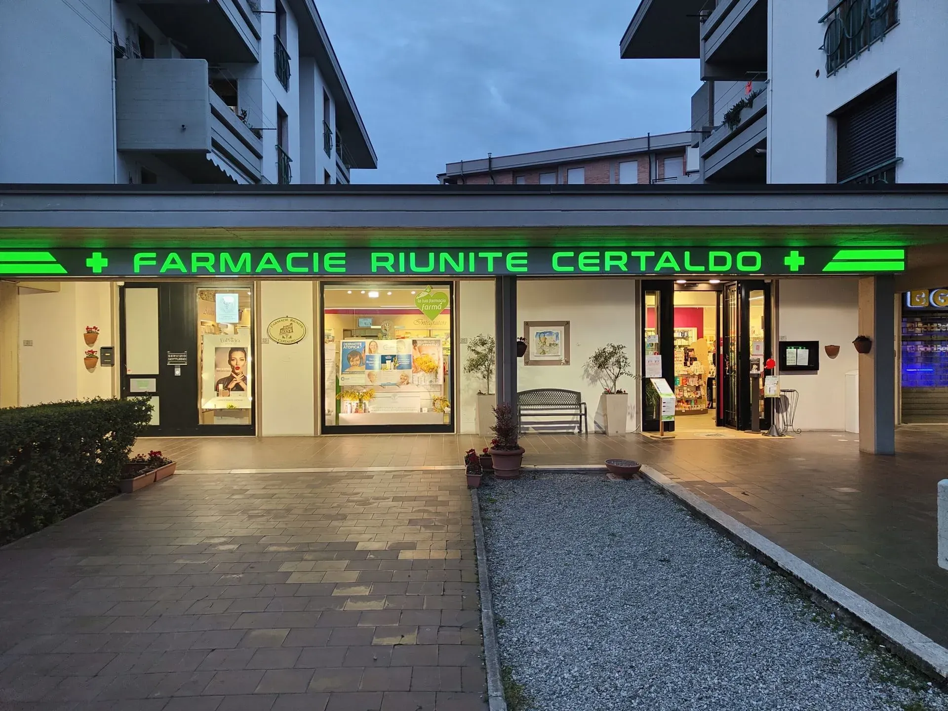 Ingresso della Farmacia Riuinte Certallo al crepuscolo, con l'insegna verde brillante e le vetrine illuminate.