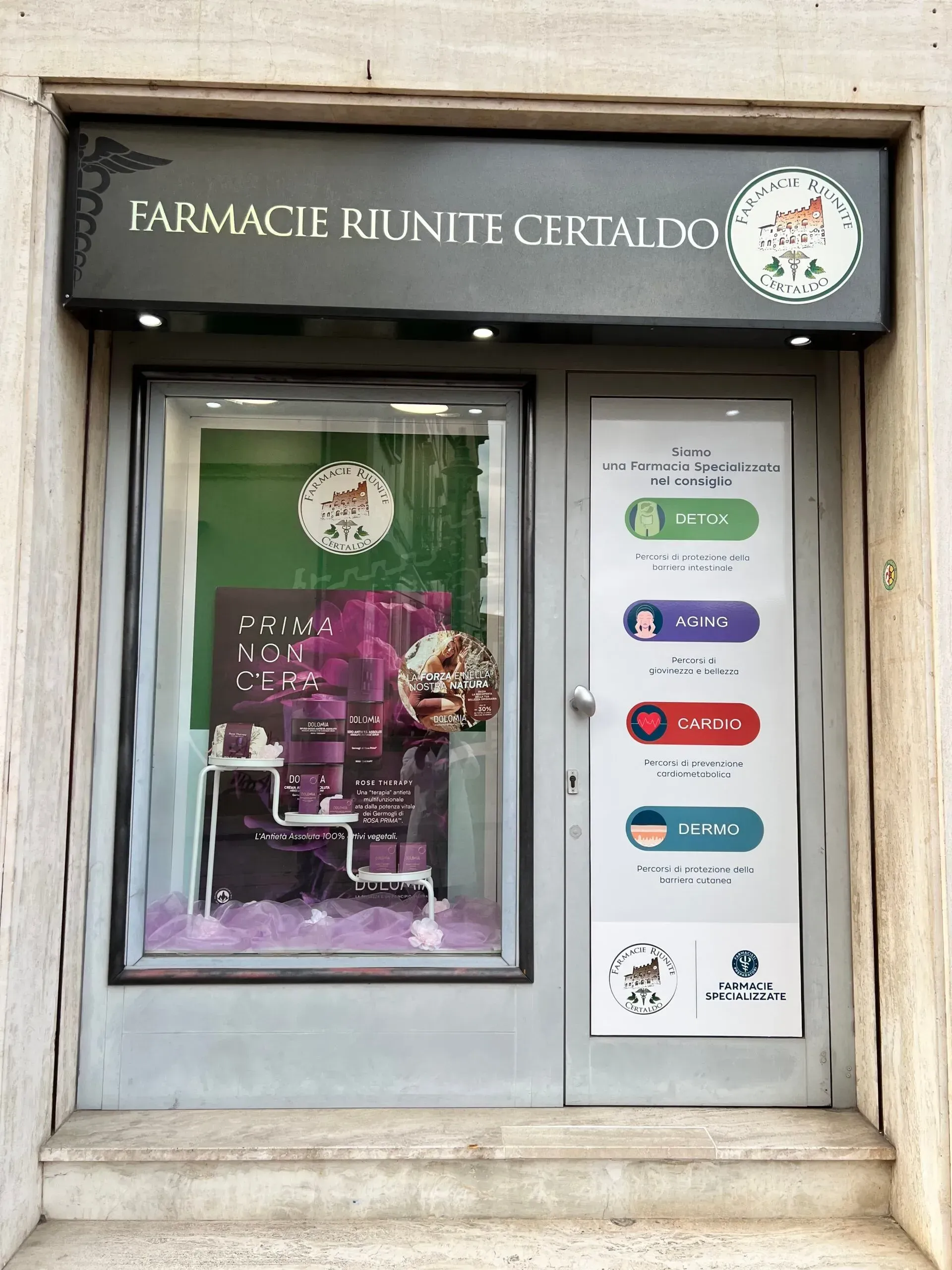 Vetrina della farmacia con porta e finestra in vetro, facciata bianca e insegna verde sopra l'ingresso.
