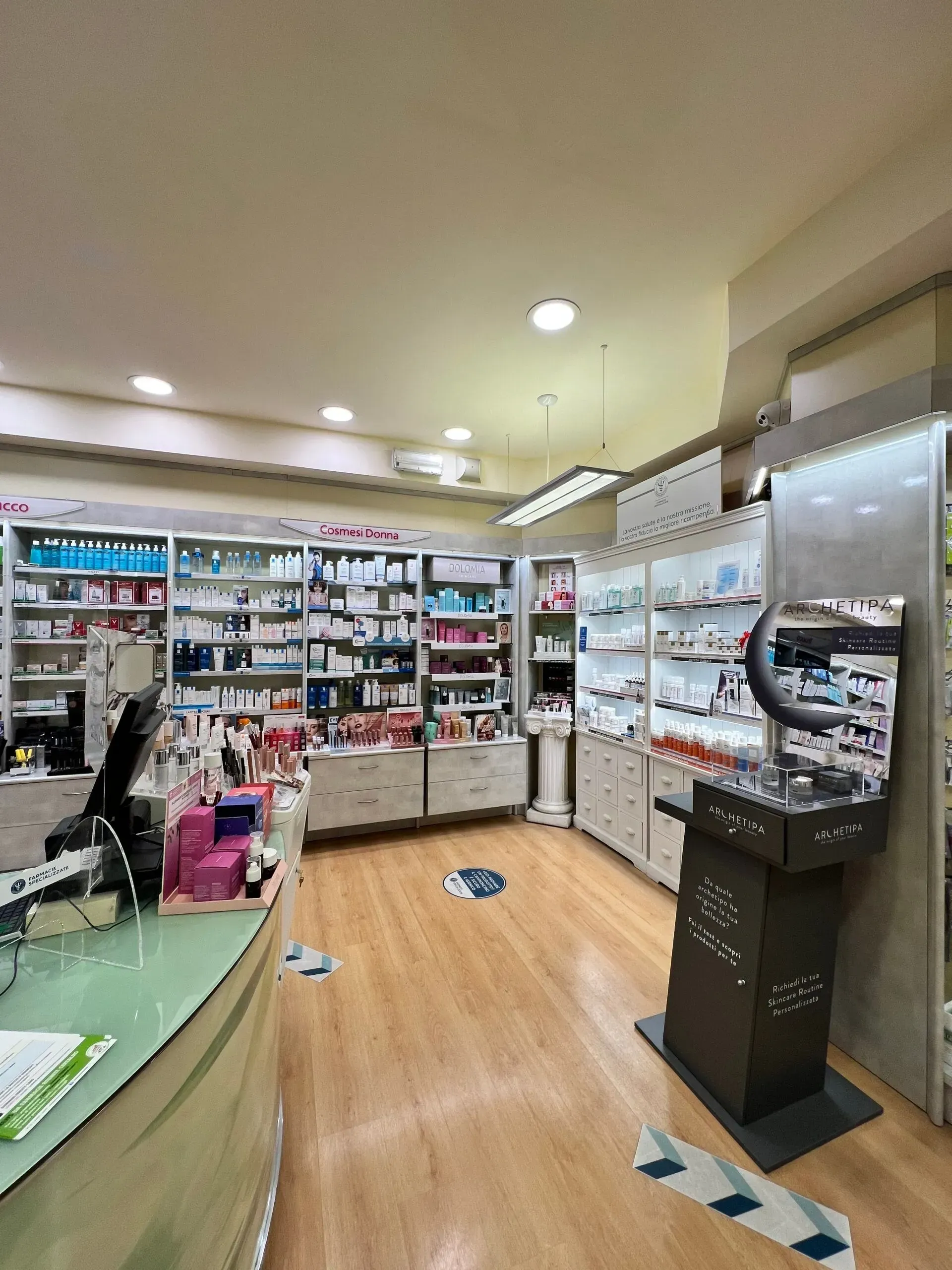 Luminosa corsia della farmacia con scaffali pieni di medicinali, un bancone e un chiosco self-service sotto calde luci del soffitto.