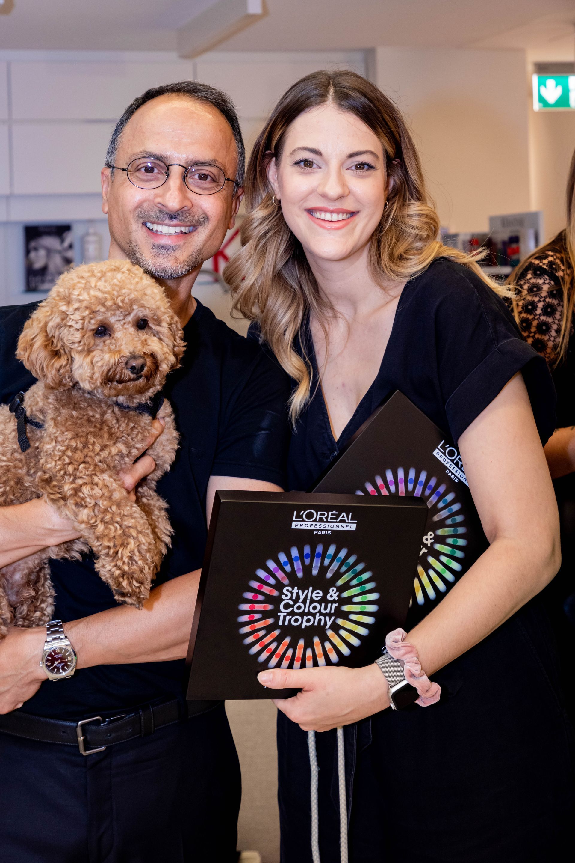 Ein Mann hält einen Pudel im Arm, eine Frau mit Aktenordnern, beide lächeln. Die Szene spielt sich in einem Innenraum ab.