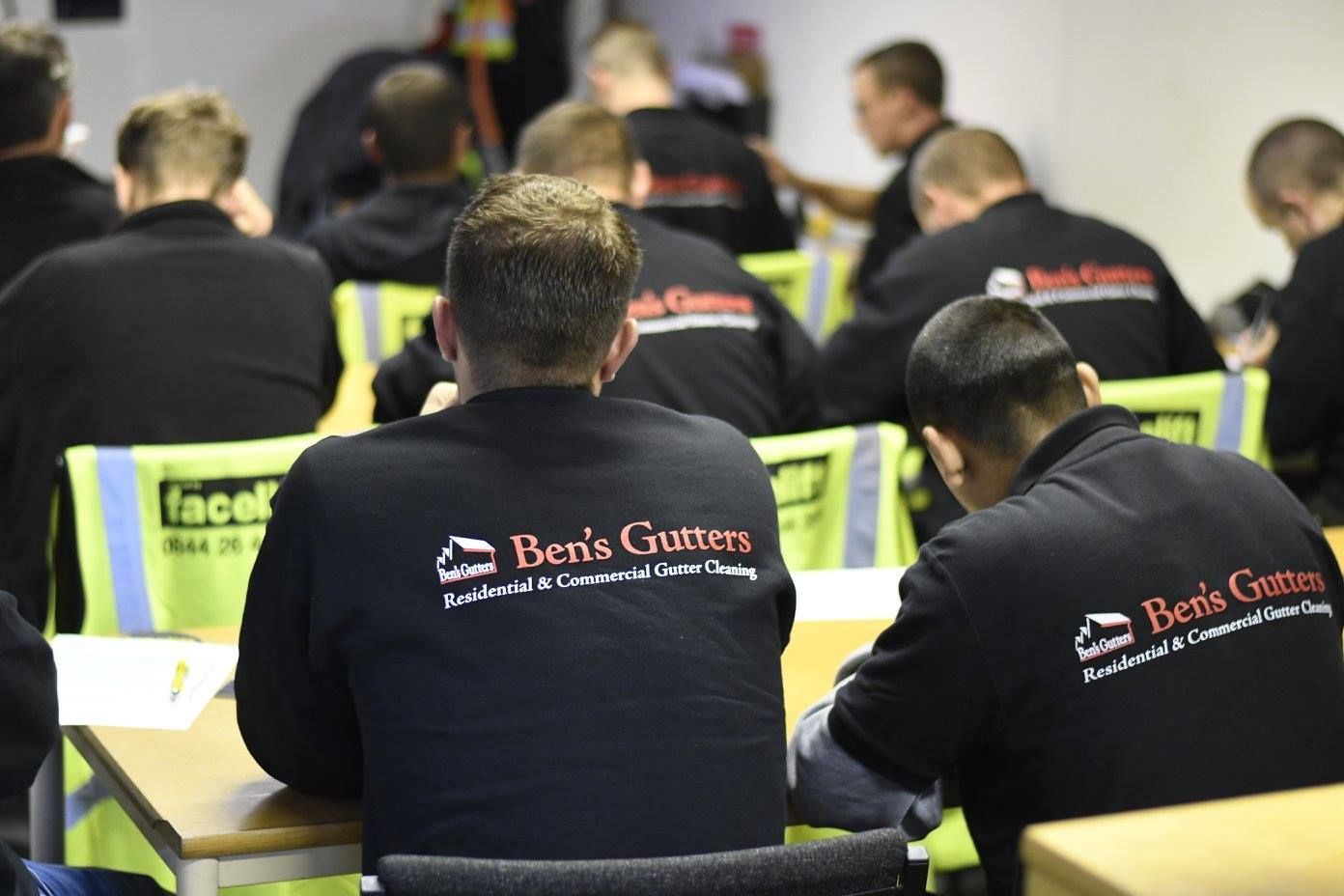People seated in a training room, seen from behind, wearing black jackets with logos.