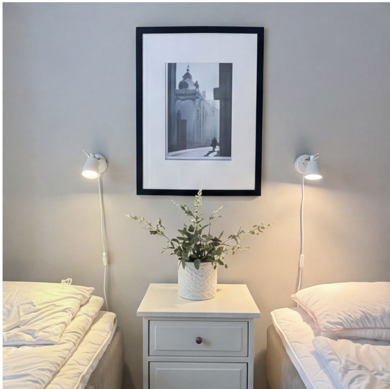 Hemtrevliga detaljer i ett dubbelrum. Två sängar med vita lakan, ett nattduksbord med en liten växt, mysiga vägglampor och en inramad tavla.
