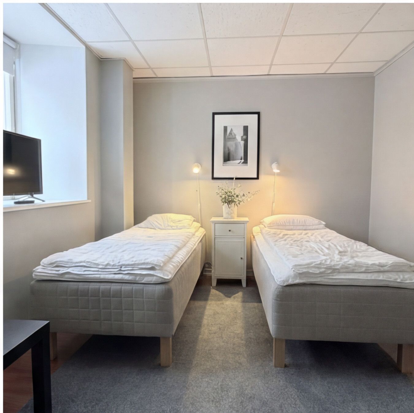 Ljust dubbelrum med två enkelsängar, nattduksbord och en tv vid fönstret.