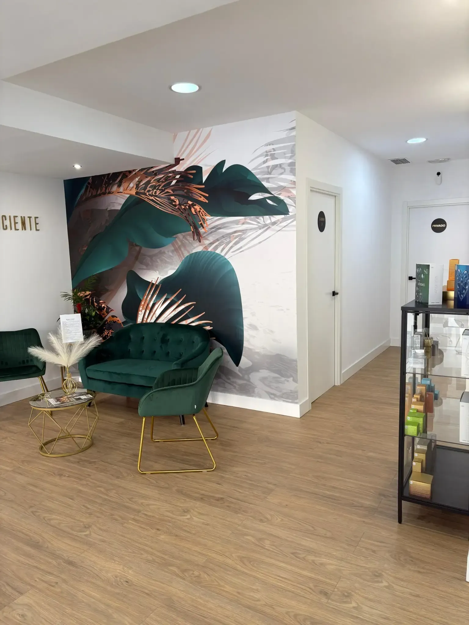 Una elegante sala de espera con una silla verde, un mural floral, detalles dorados y exhibición de productos.