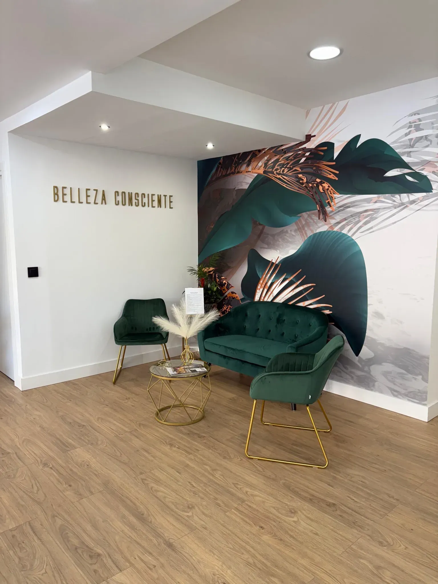 Área de espera de un salón de belleza con asientos verde esmeralda, detalles dorados y un mural botánico.