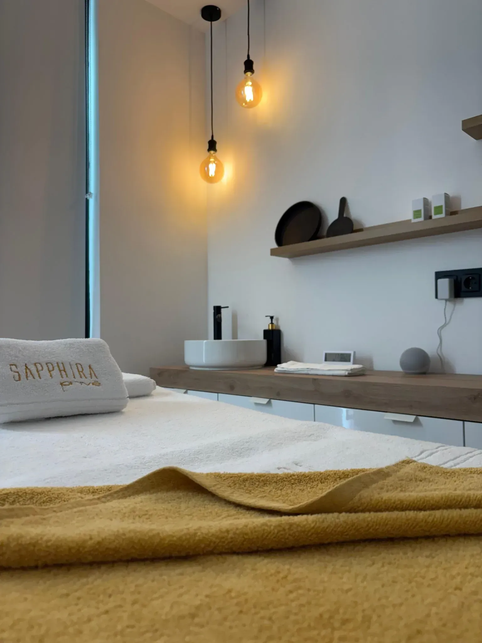 Sala de spa con cama de masajes, luces de techo, estanterías de madera y paredes blancas.