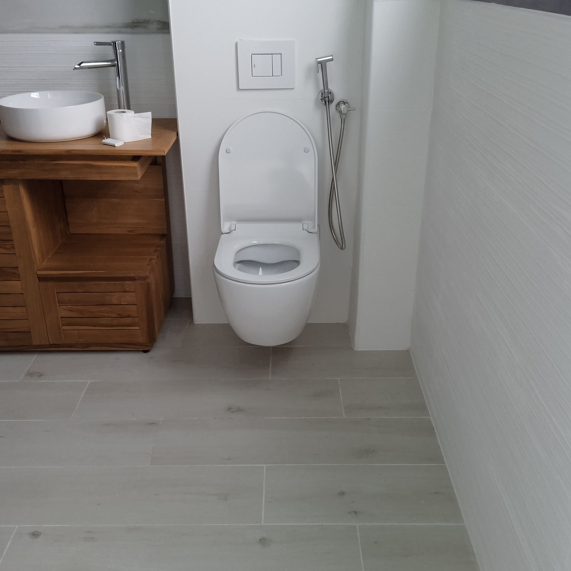 Petite salle de bains : toilettes suspendues avec douchette chromée sr la droite, meuble lavabo en bois avec vasque blanche.