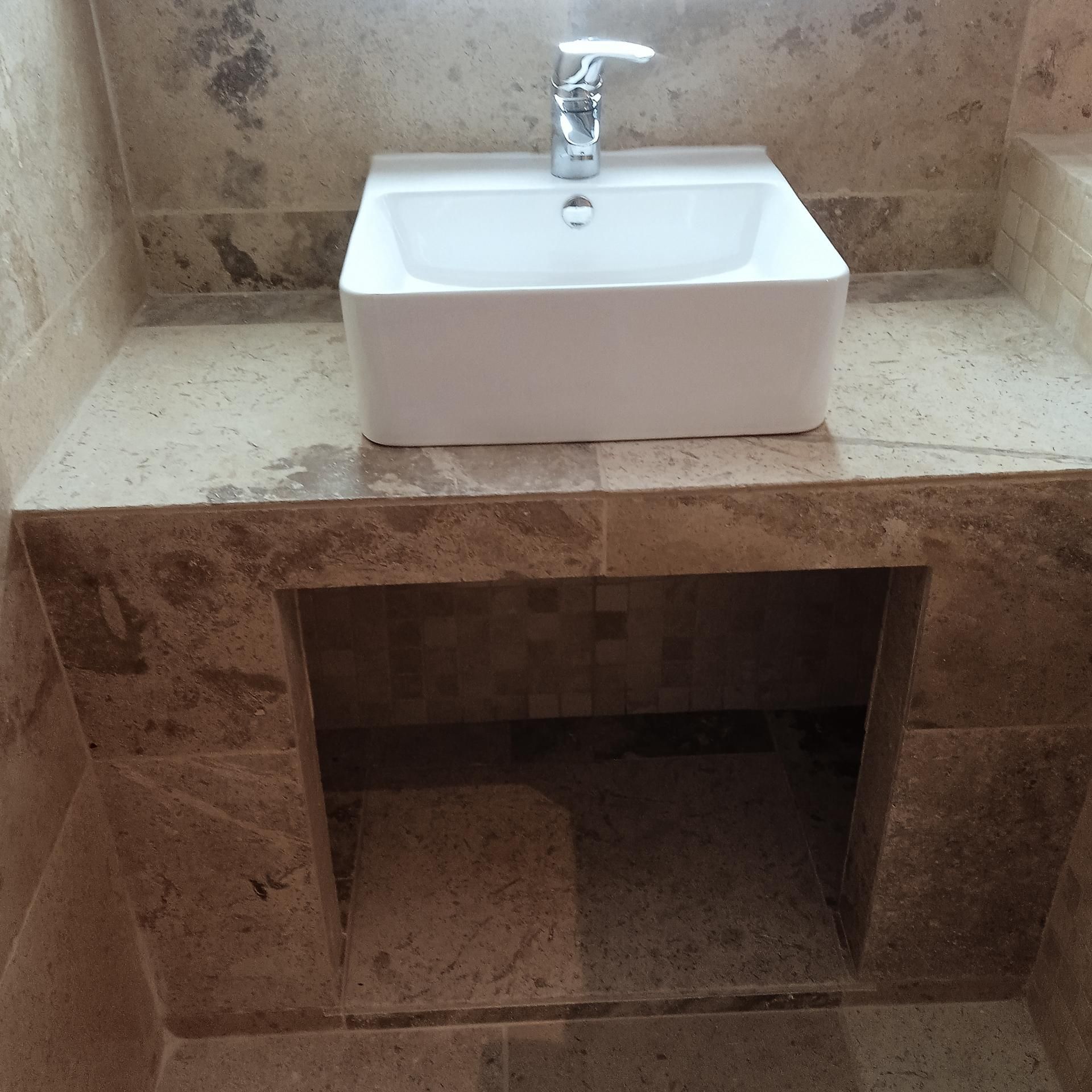 Lavabo blanc avec robinet chromé sur un comptoir de salle de bains en pierre, au-dessus d'un espace de rangement ouvert.