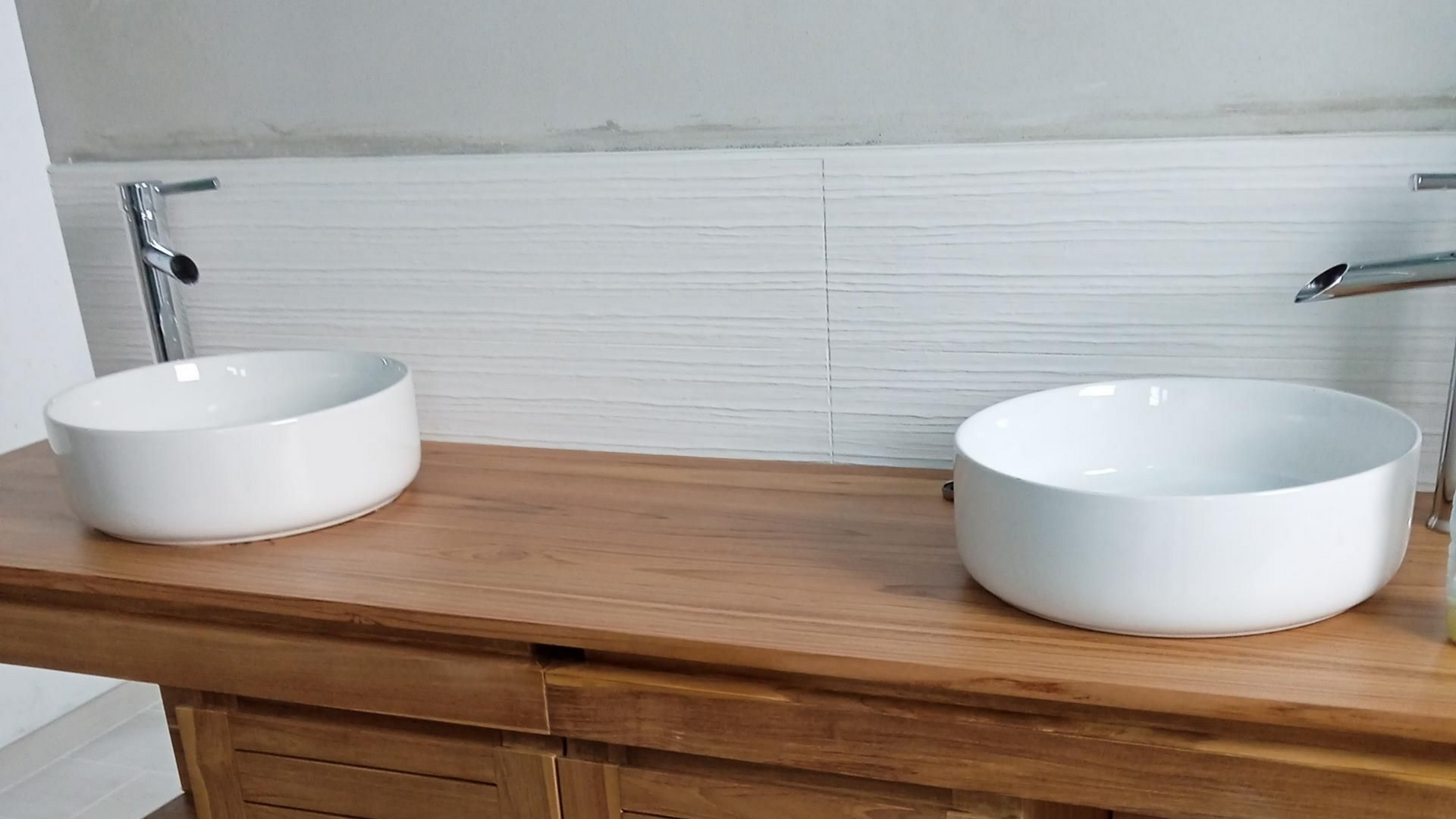 Deux lavabos ronds blancs sur un plan de travail en bois, devant un mur carrelé clair.