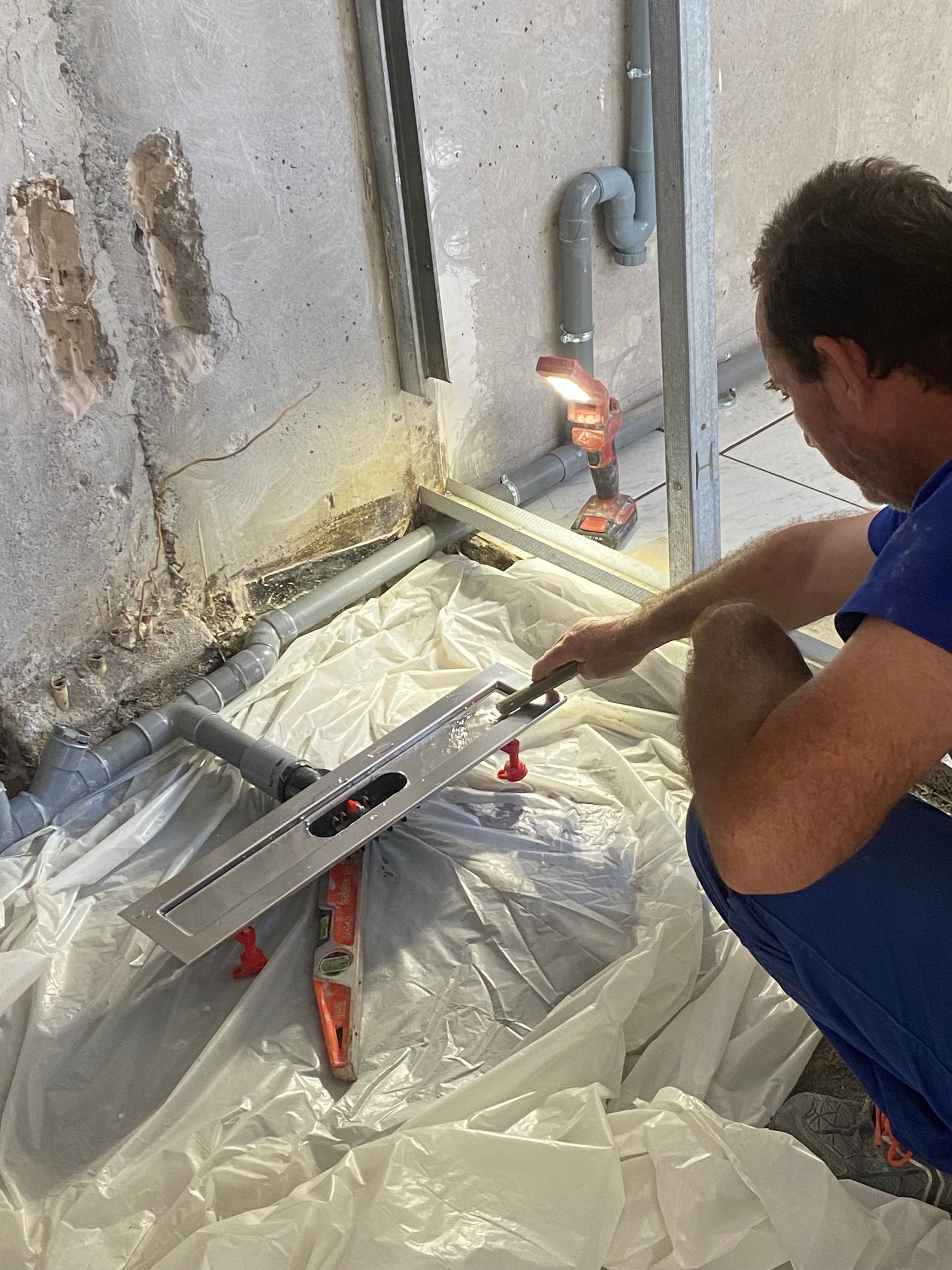 Un ouvrier, agenouillé sur une bâche en plastique, mesure et nivelle un sol près d'un mur en construction.