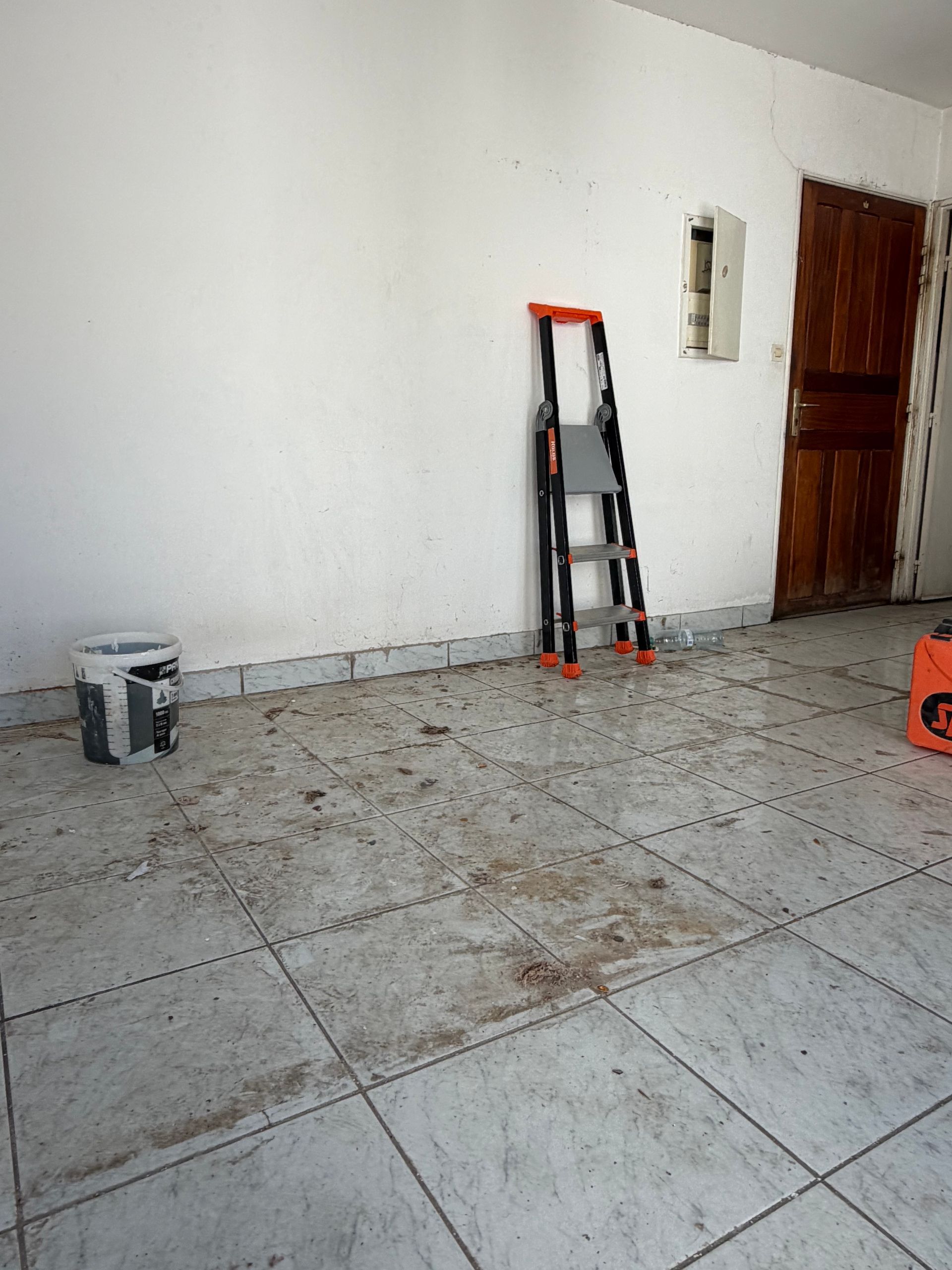 Pièce spartiate avec sol carrelé, échelle appuyée contre le mur et porte en bois sur la droite.