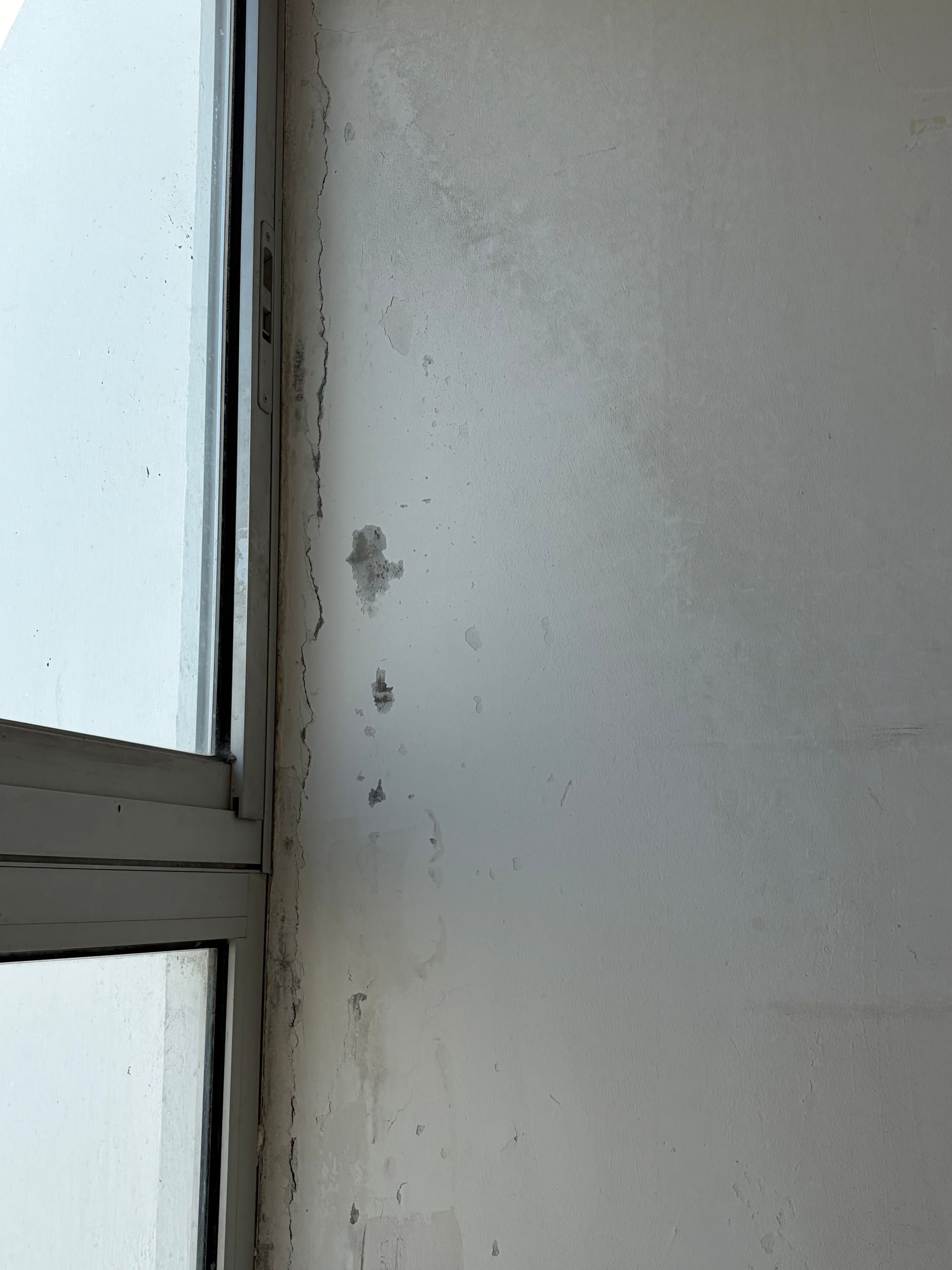 Gros plan sur un mur blanc à côté d'un cadre de fenêtre présentant de petites taches et des zones de peinture écaillée.