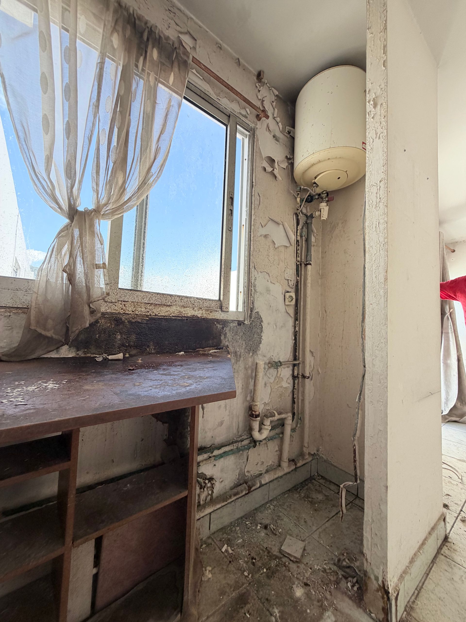 Cuisine poussiéreuse et abîmée, avec une fenêtre cassée, un chauffe-eau mural et une armoire en bois remplie de débris.