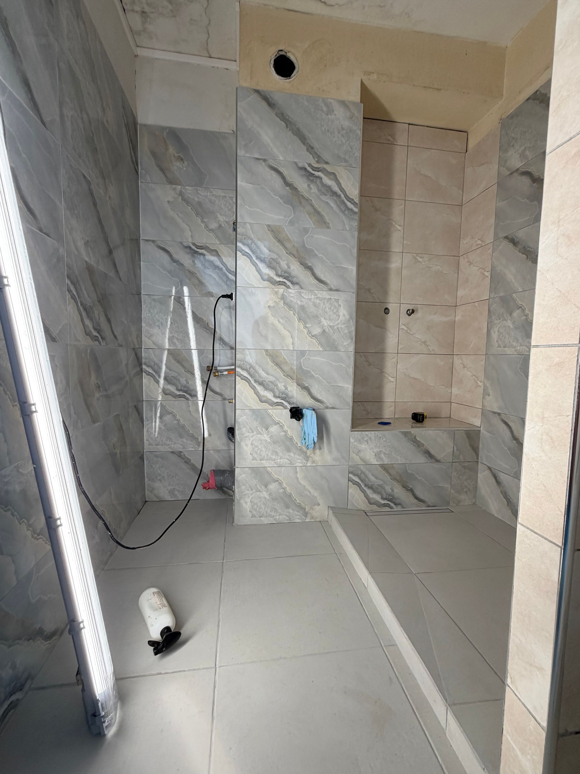 Salle de bains carrelée en cours de construction avec murs imitation marbre, robinetterie de douche et outils au sol.