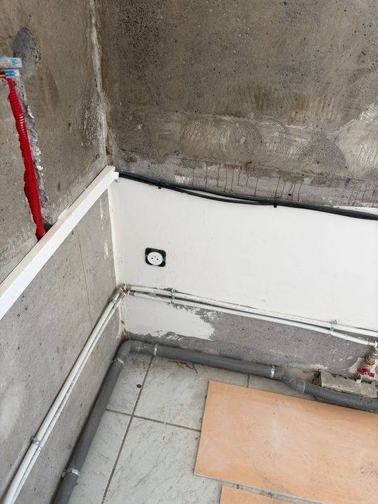 Angle de mur brut en béton avec traces d’humidité, gaine électrique rouge, prise murale et tuyaux apparents au sol.