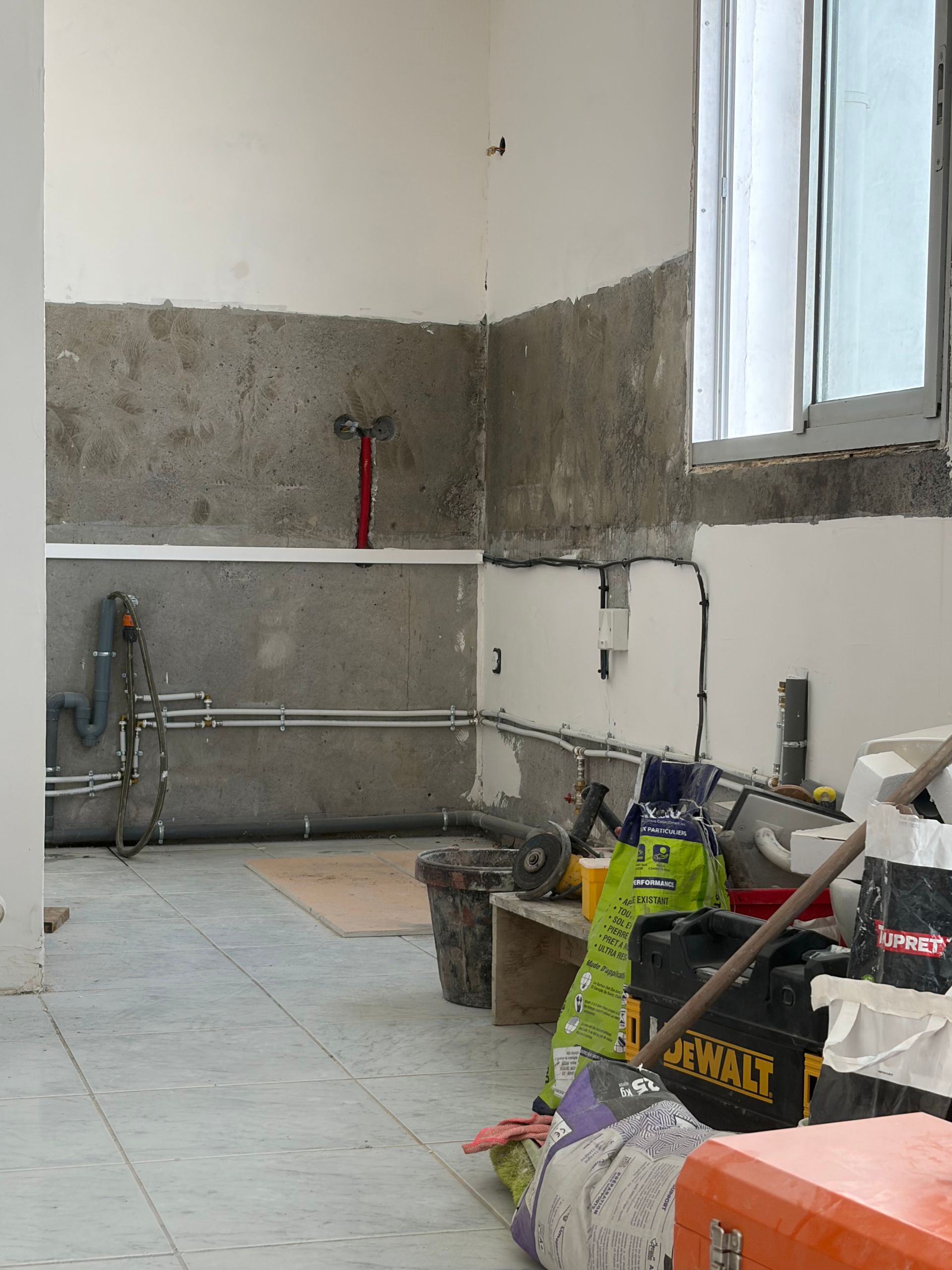 Pièce en rénovation avec murs inachevés, outils, seaux et matériaux de construction près d'une fenêtre.