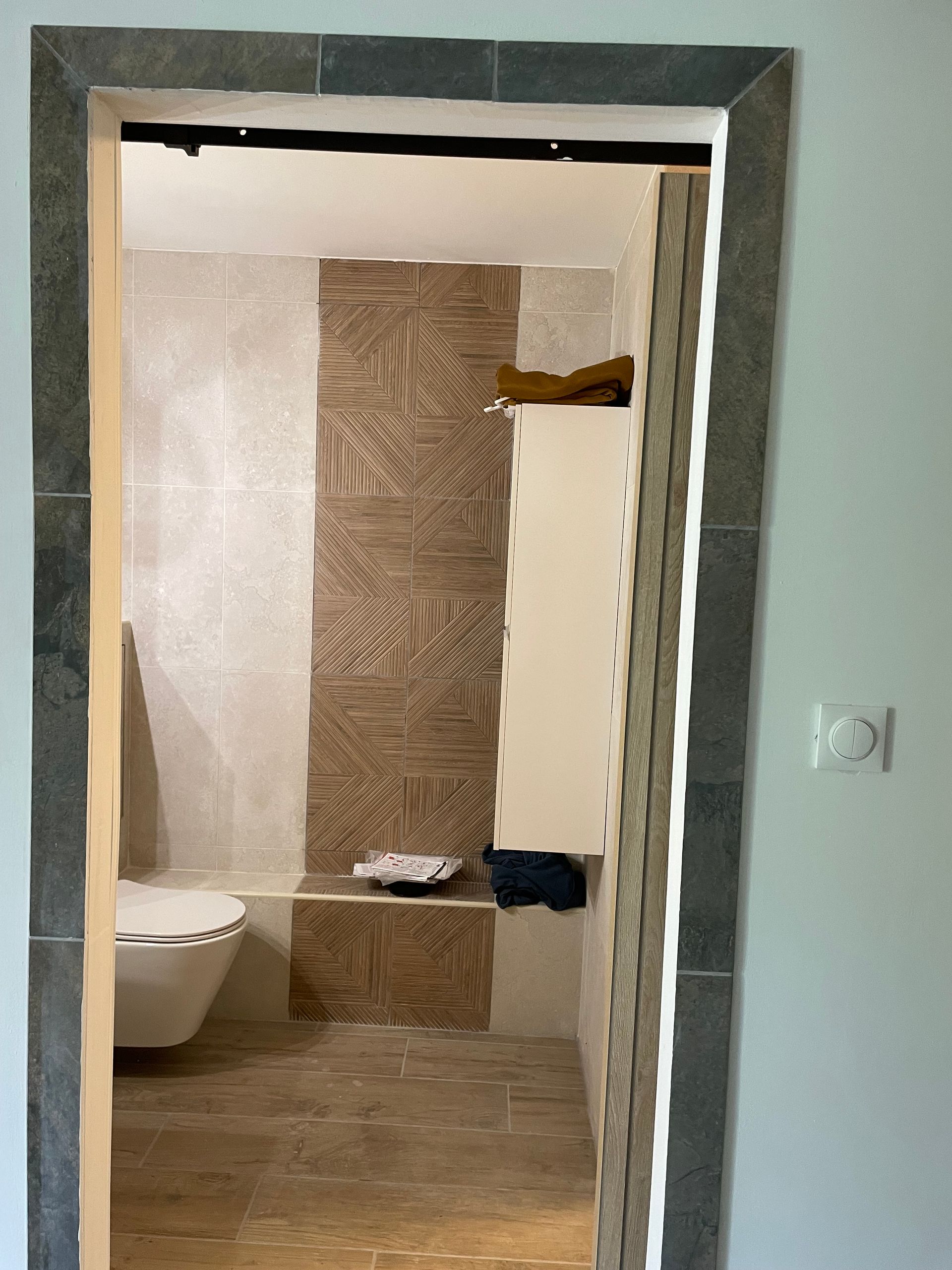 Vue de la porte de la salle de bains : toilettes suspendues, carrelage mural gris et revêtement à motifs imitation bois.
