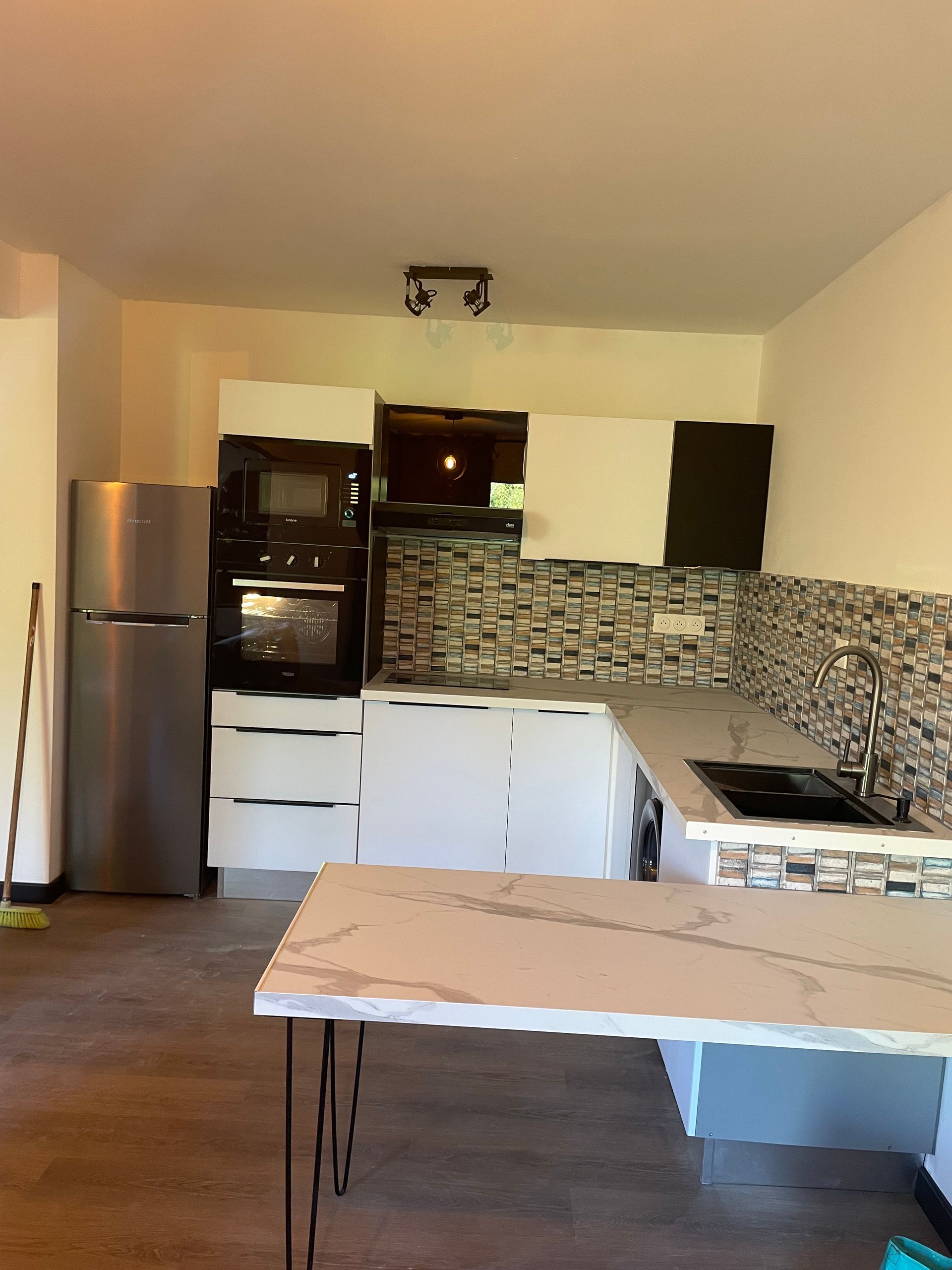 Petite cuisine moderne avec réfrigérateur en inox, placards blancs, crédence en mosaïque et électroménager noir.