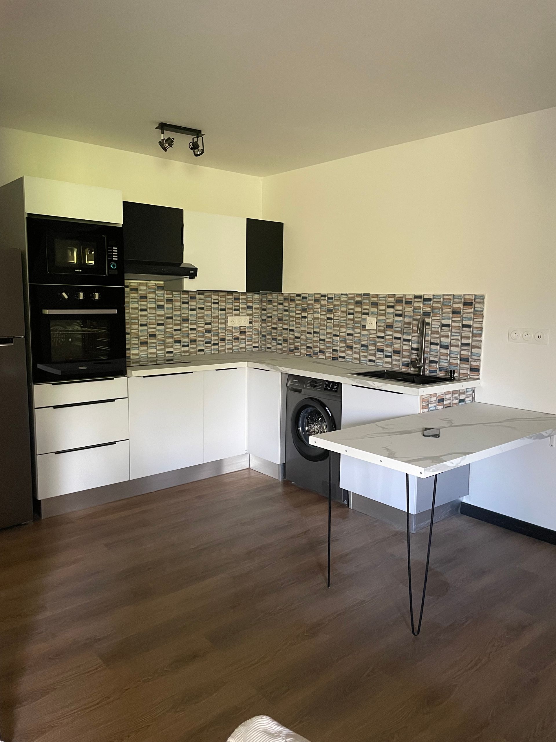 Cuisine moderne avec armoires blanches, appareils électroménagers noirs, crédence en mosaïque et petite table au premier plan.