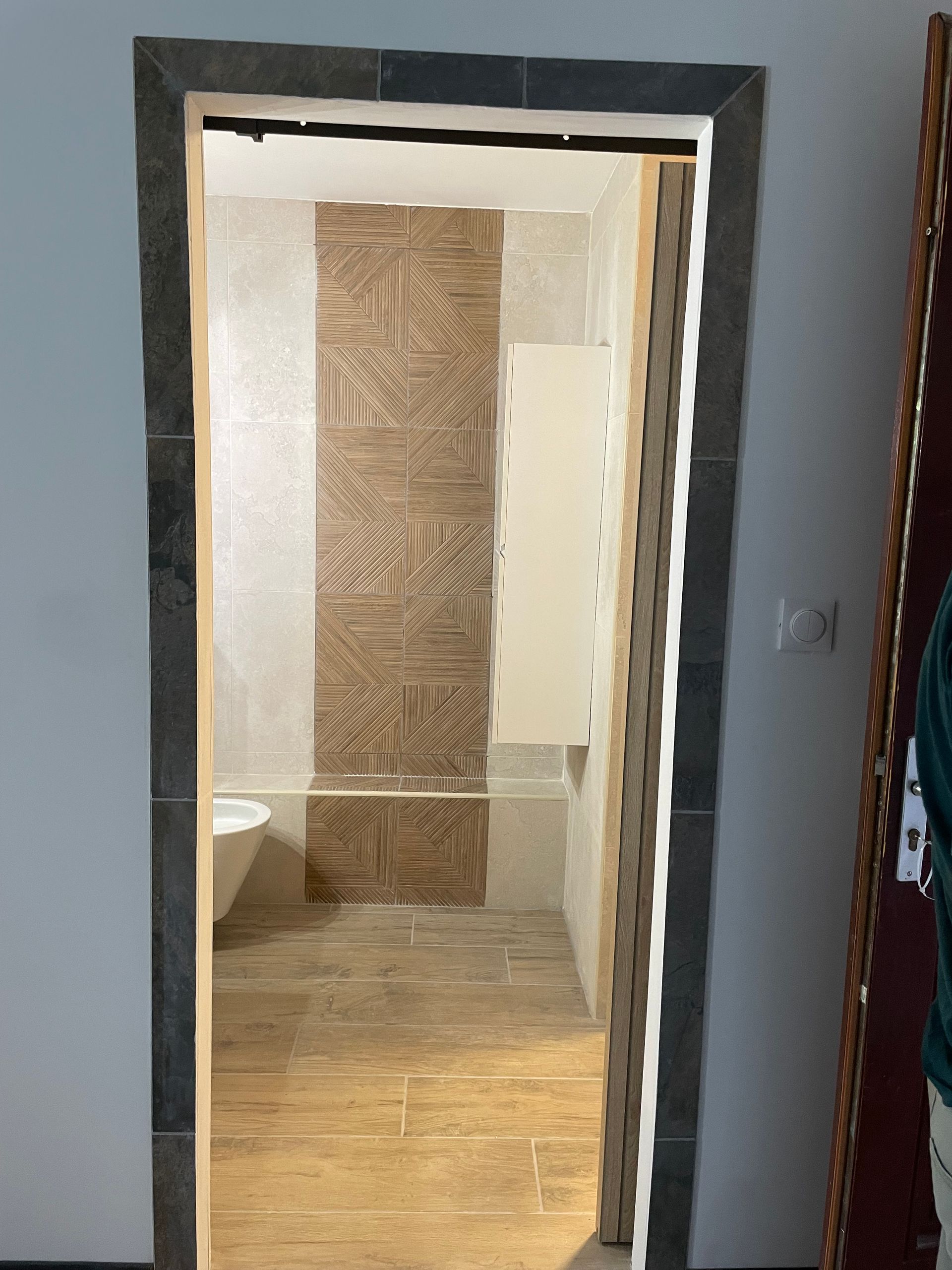 Porte de salle de bains ouverte avec vue sur des toilettes suspendues, un carrelage gris et un revêtement imitation bois.