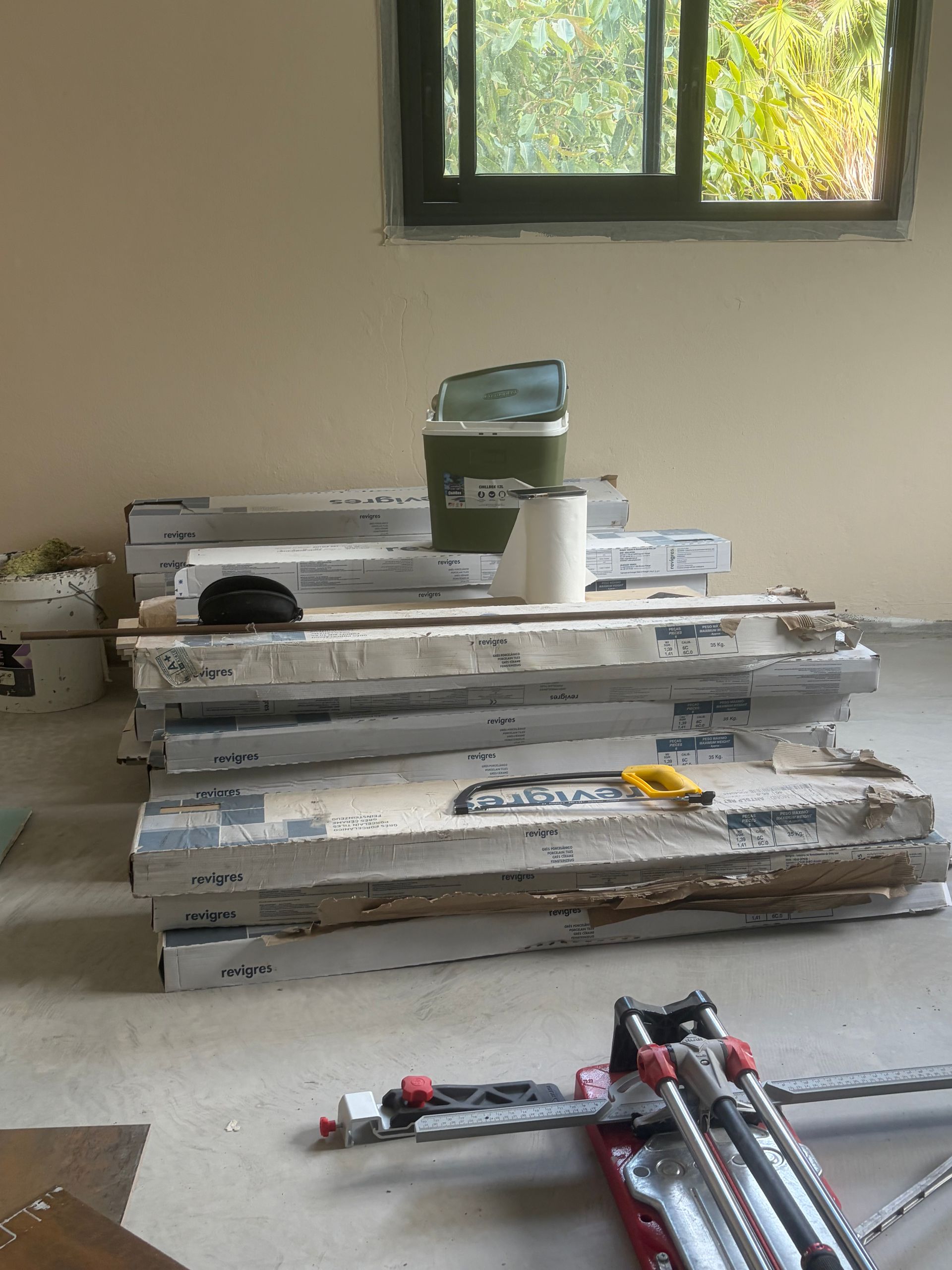 Des cartons de carrelage en grès cérame posés dans une pièce en rénovation, avec un trépied plié et une fenêtre ouverte.