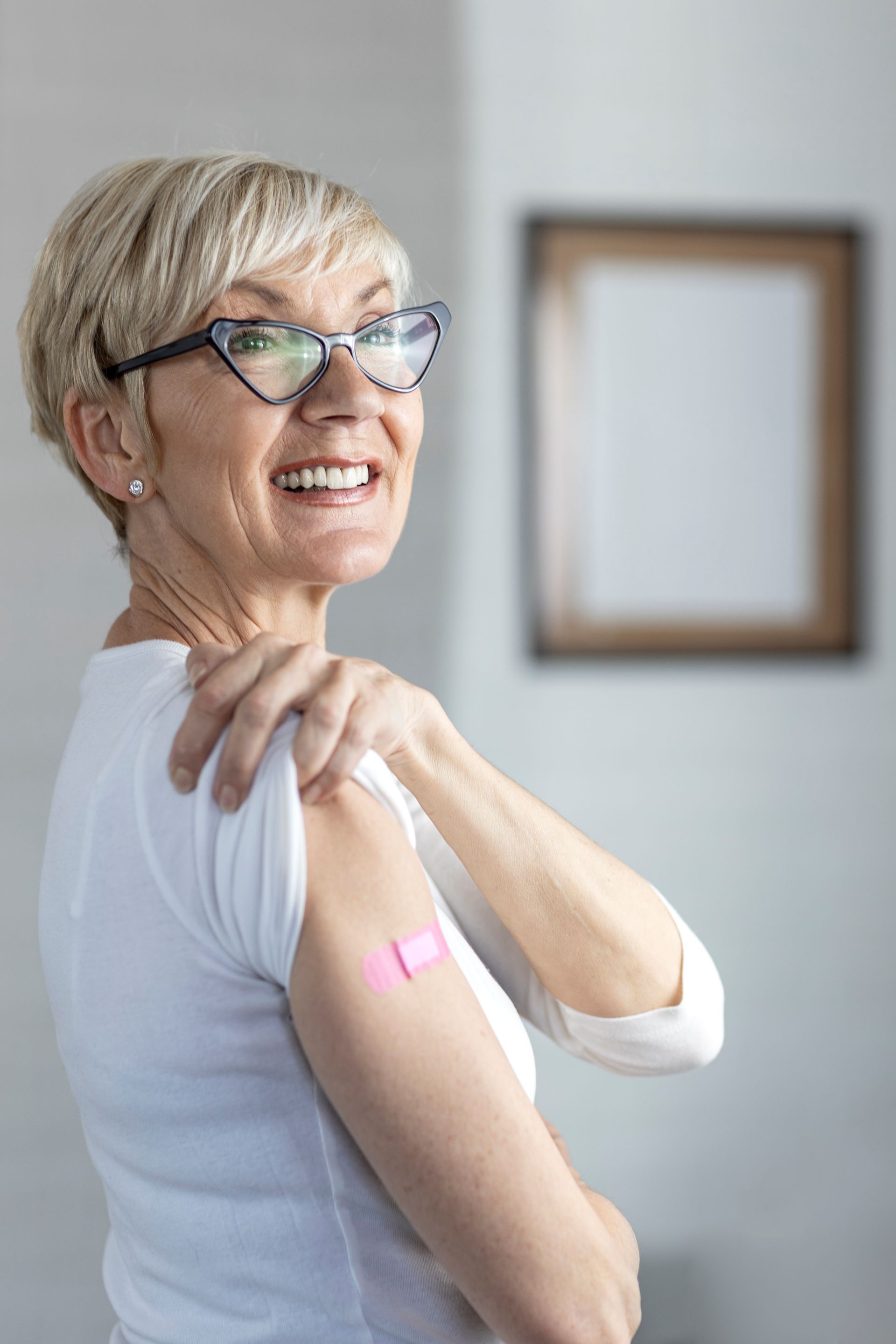 Une femme portant des lunettes sourit, tenant un bras avec un bandage, probablement vaccinée, dans une pièce avec une œuvre d'art encadrée.