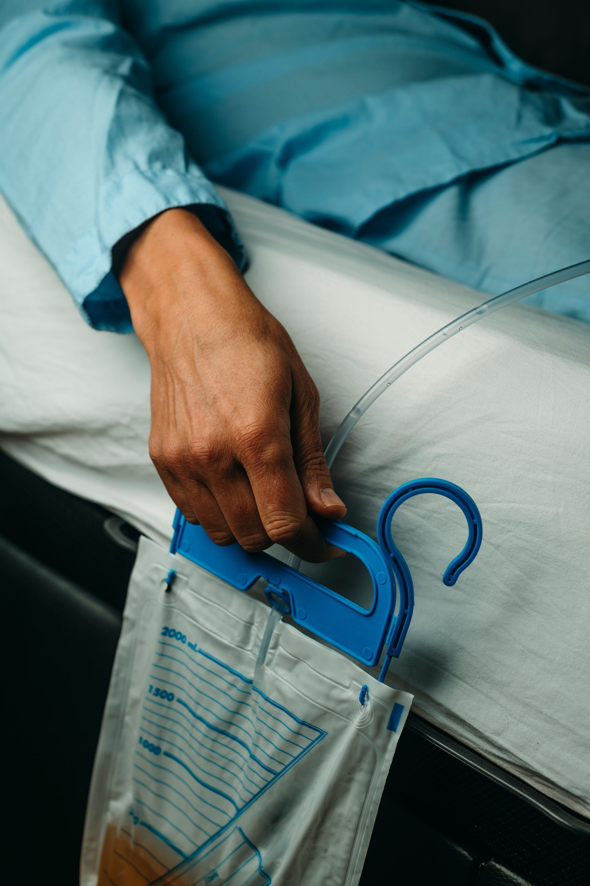 Main d'une personne tenant un sac de collecte d'urine à poignée bleue relié à un tube, sur un lit d'hôpital.