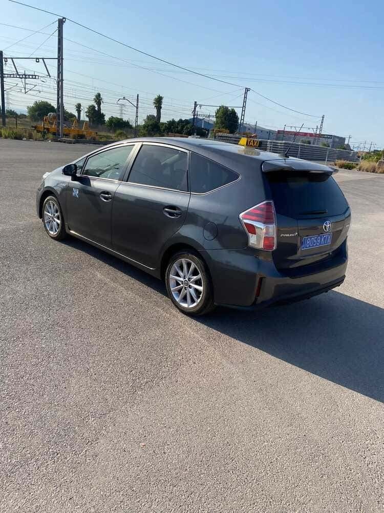 Toyota Prius V gris oscuro estacionado sobre asfalto con vías de tren al fondo.