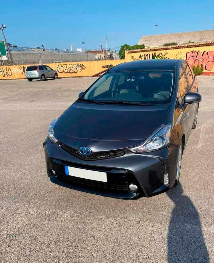 Toyota Prius híbrido gris oscuro estacionado en un estacionamiento, con un cielo azul y una pared amarilla en el fondo.