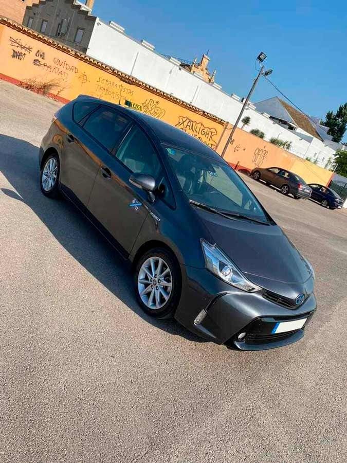 Toyota Prius V gris oscuro estacionado sobre asfalto, en un día soleado. Fondo: pared de grafitis y otros coches.
