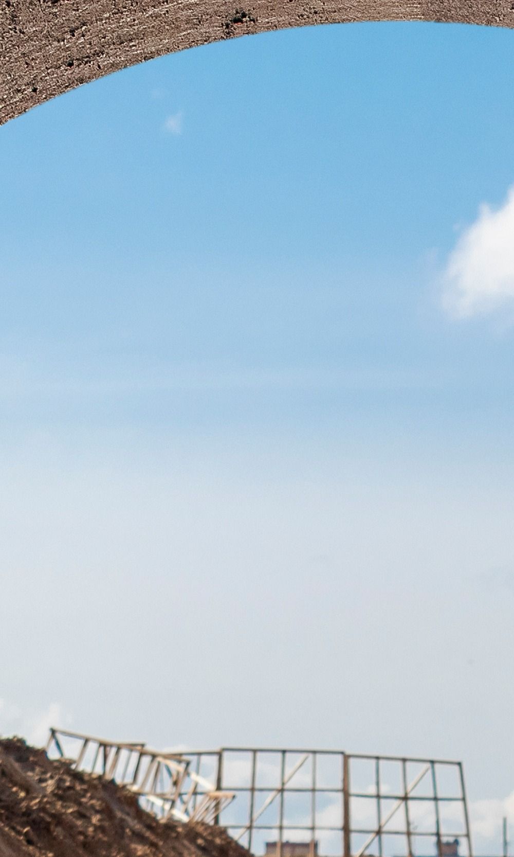 Ciel bleu parsemé de nuages épars, visible à travers une arche de matière brune ressemblant à du gravier ; chantier en contrebas.