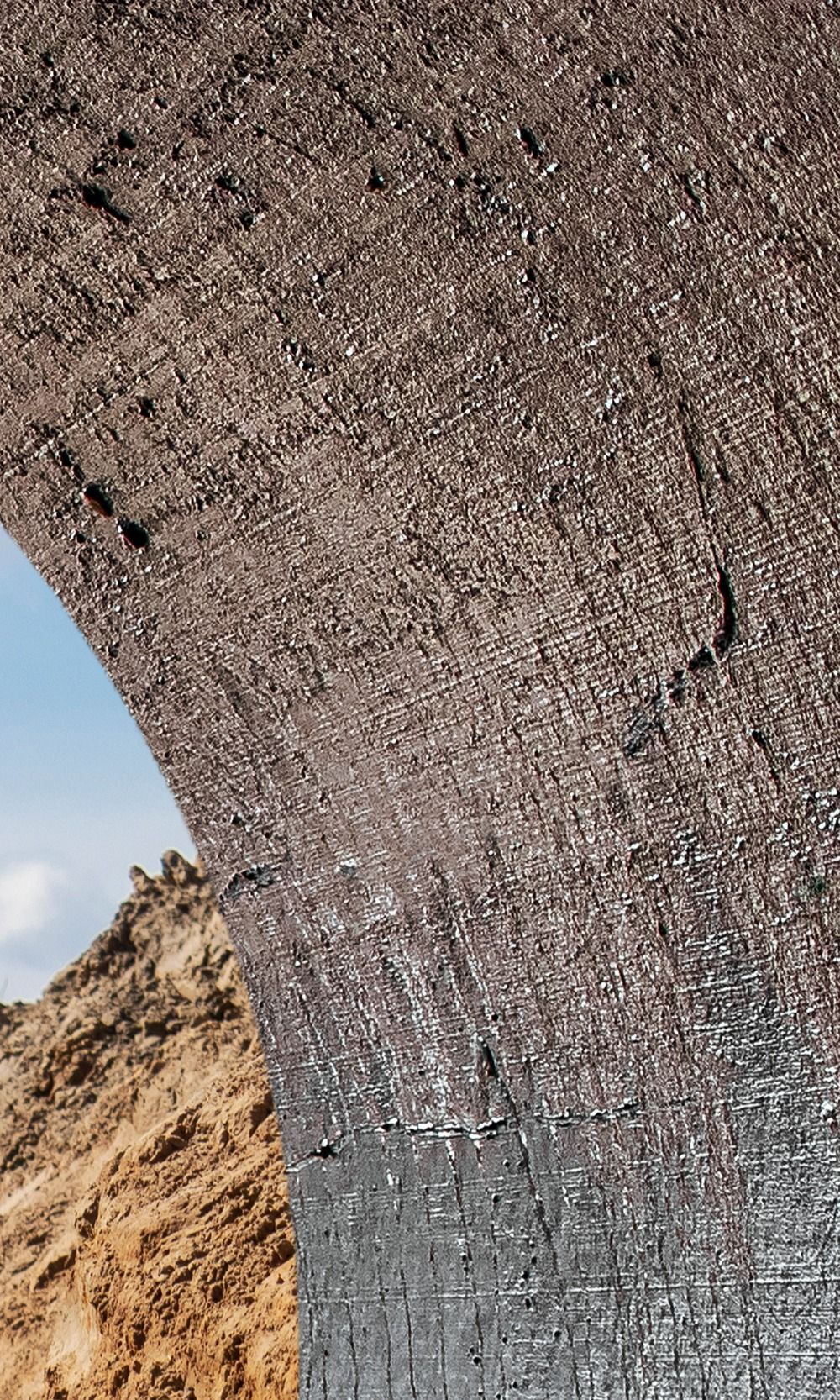 Gros plan sur une surface texturée et arquée, probablement de la roche ou du béton, avec un aperçu de ciel bleu et d'un paysage beige dans l'ouverture.