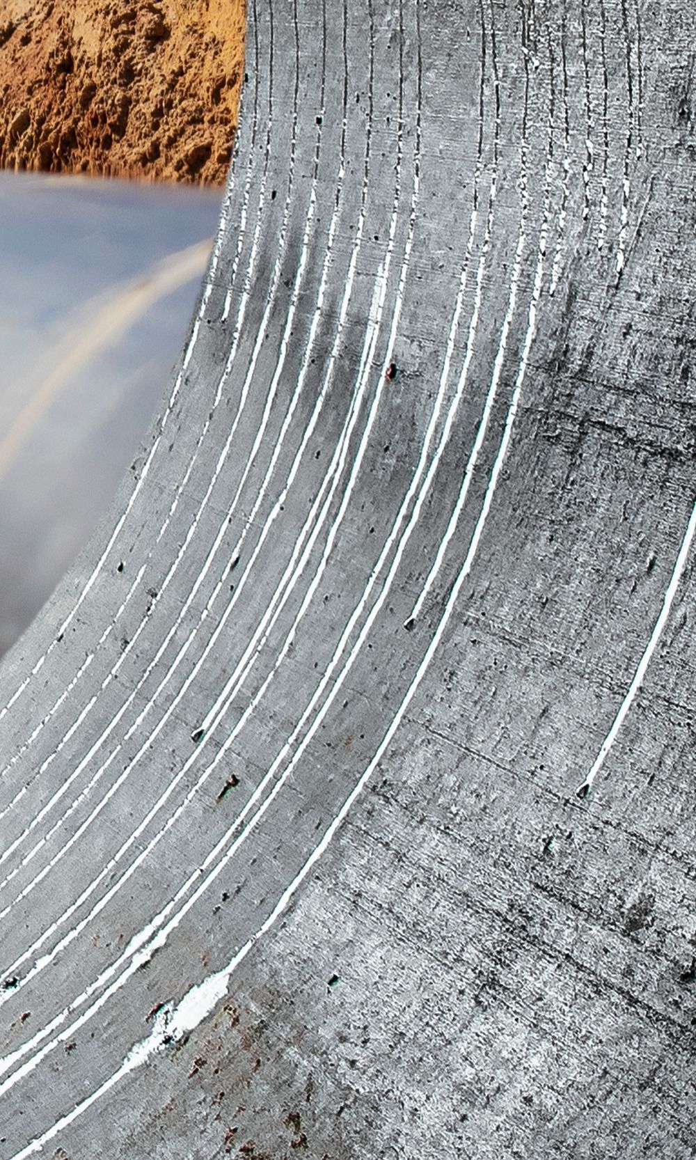 Surface incurvée en béton gris avec de longues stries blanches, probablement de la peinture ou des résidus. Terre en arrière-plan.