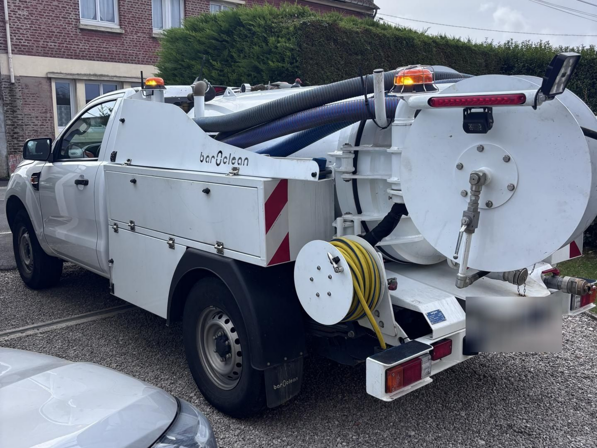 Camion blanc avec réservoir et tuyaux, stationné sur une surface pavée près d'un bâtiment.