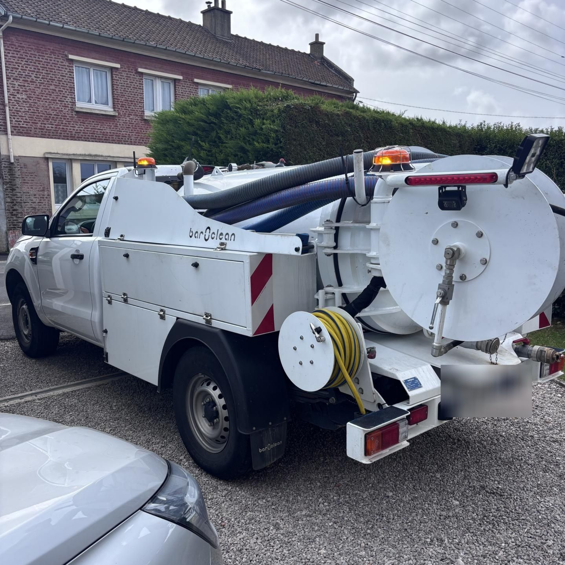 Camion aspirateur blanc stationné sur une allée de gravier devant une maison en briques.