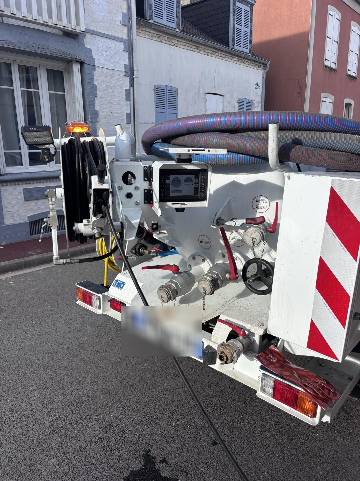 Vue arrière d'un camion utilitaire blanc dans une rue, montrant divers équipements et tuyaux. Panneau à rayures rouges et blanches visible.