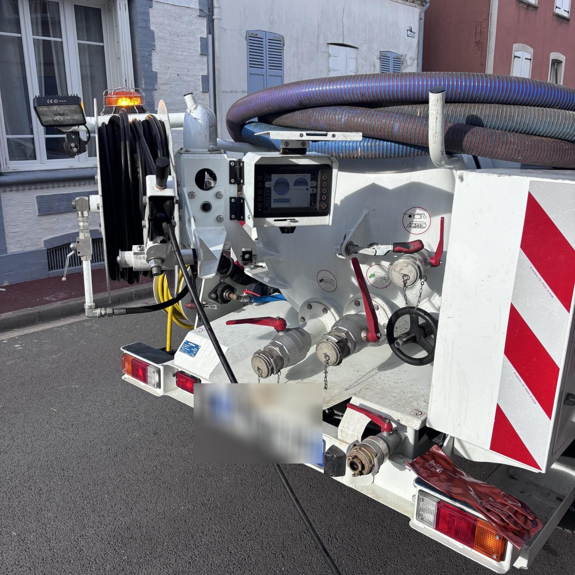 Vue arrière d'un camion de service blanc avec tuyaux, panneau de commande et feux de signalisation, stationné dans une rue.