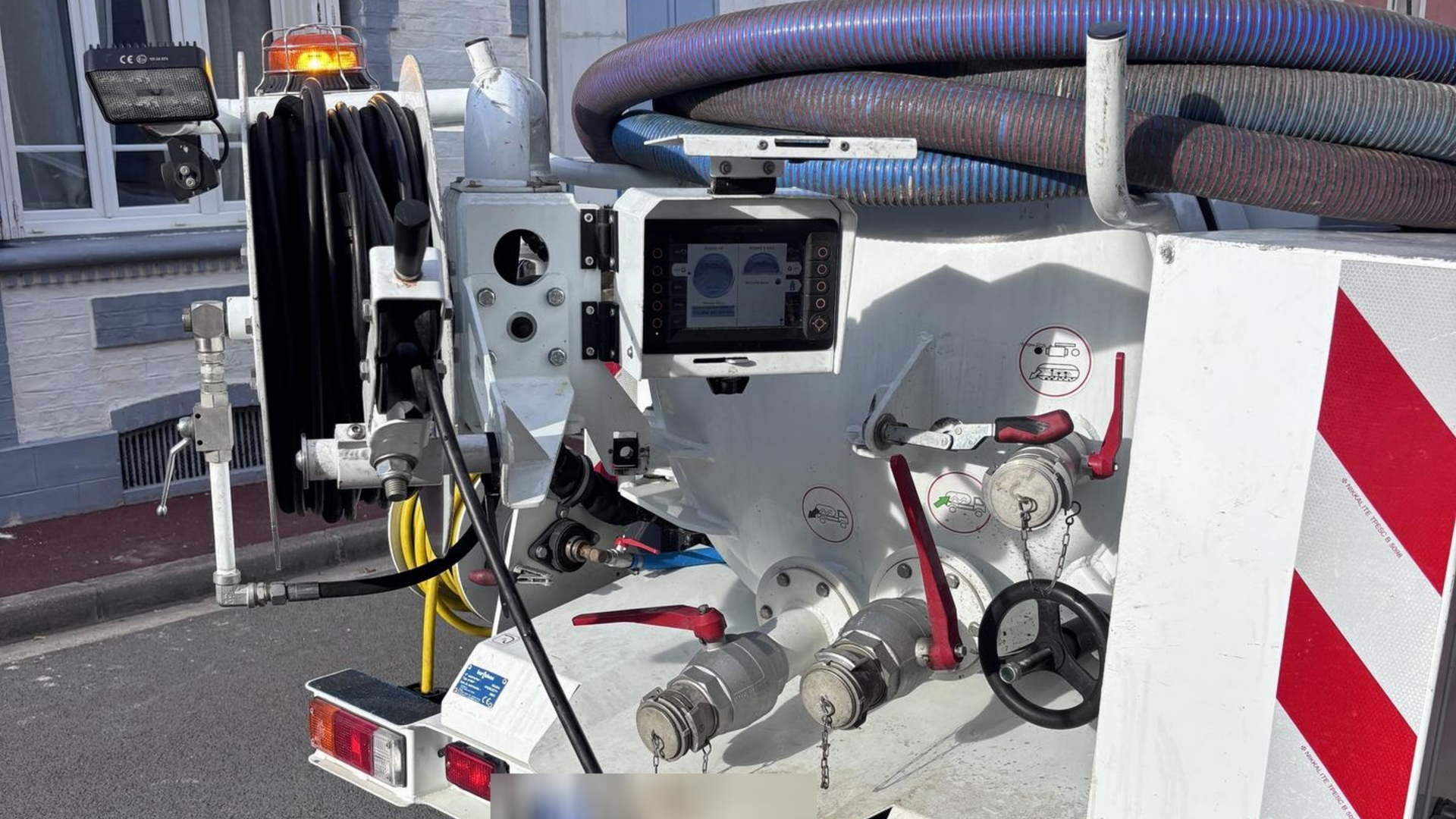 Panneau de commande d'un camion blanc avec tuyaux et écran, en bordure de rue. Feux de signalisation et bandes réfléchissantes présents.